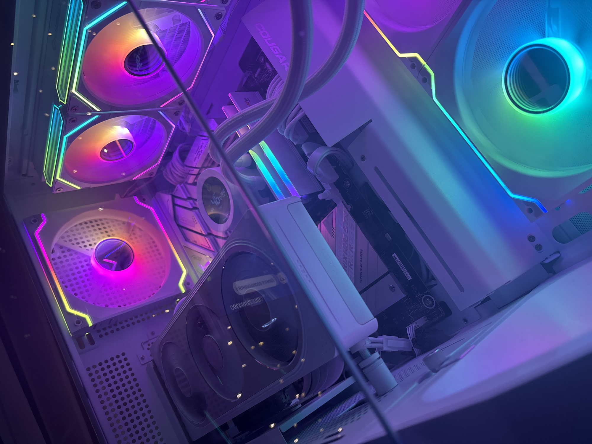 A close-up, high-angle shot captures the interior of a white computer tower case illuminated by an array of colorful RGB lights. The lighting shifts through shades of purple, pink, yellow, green, and blue, creating a vibrant and futuristic aesthetic. Prominently featured are several large computer fans with illuminated rims, some angled towards the viewer, others oriented to move air. A white liquid cooling tube snakes across the scene, connecting to a white component bearing the "COUGAR" logo. A graphics card, also with illuminated accents, is visible towards the bottom of the frame. Various internal computer components, including circuit boards and wiring, are discernible beneath the dazzling light display. The overall impression is one of a high-performance gaming or workstation computer, showcasing custom lighting and cooling solutions. The scene is set in an indoor environment, with no clear indication of a specific city or location.