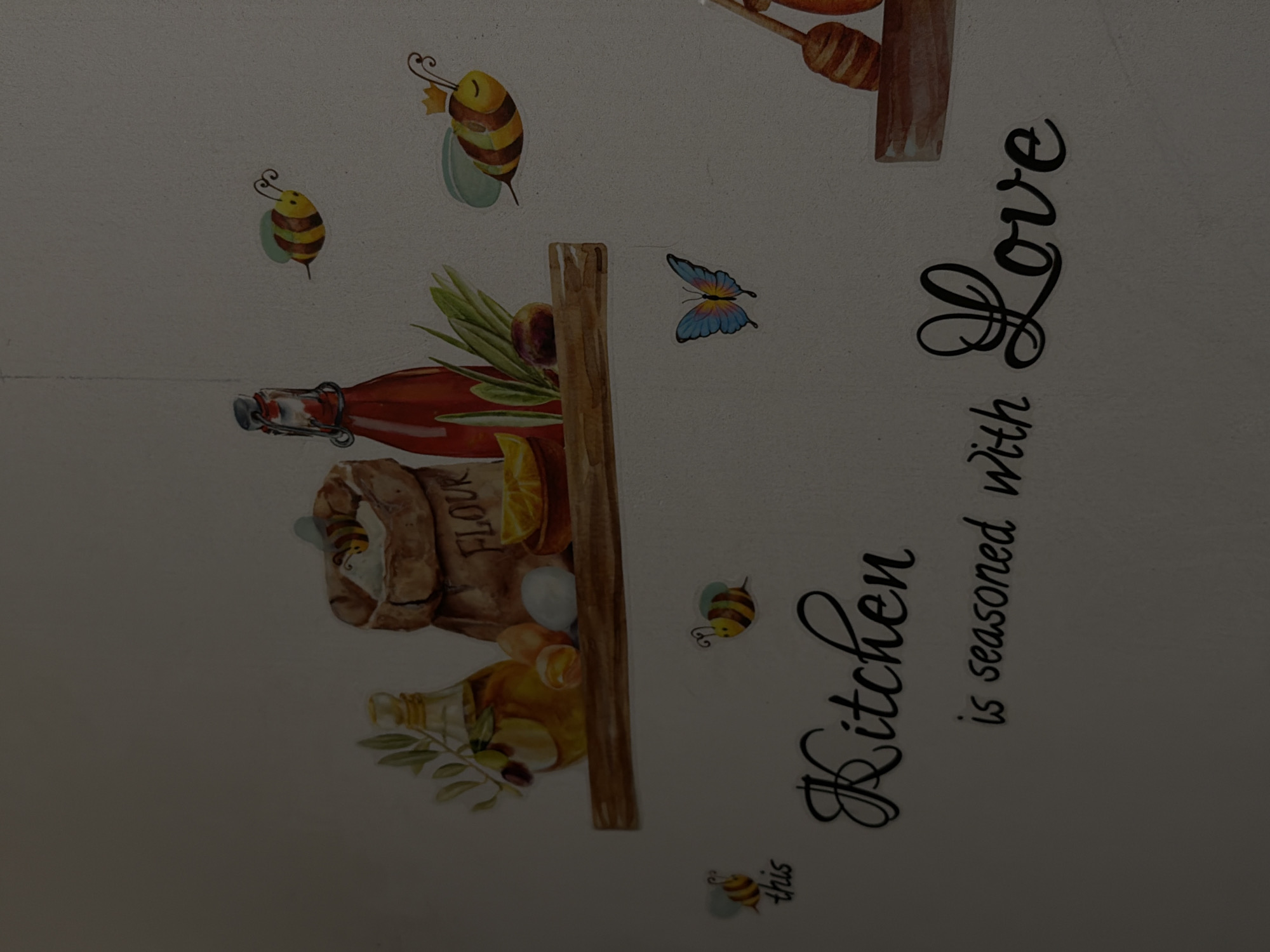 A top-down, tilted close-up shows a wall decoration with kitchen motifs. The background is a light color. Several cartoon bees are scattered about. A red bottle, a bag of "FLOUR", a halved orange, a jar of oil, and olives are depicted along with other objects on a watercolor shelf. A blue butterfly is visible near the center. The words "this Kitchen is seasoned with Love" are printed in black cursive along the right side. The image has no discernible connection to Cheltuitori, Moldova.