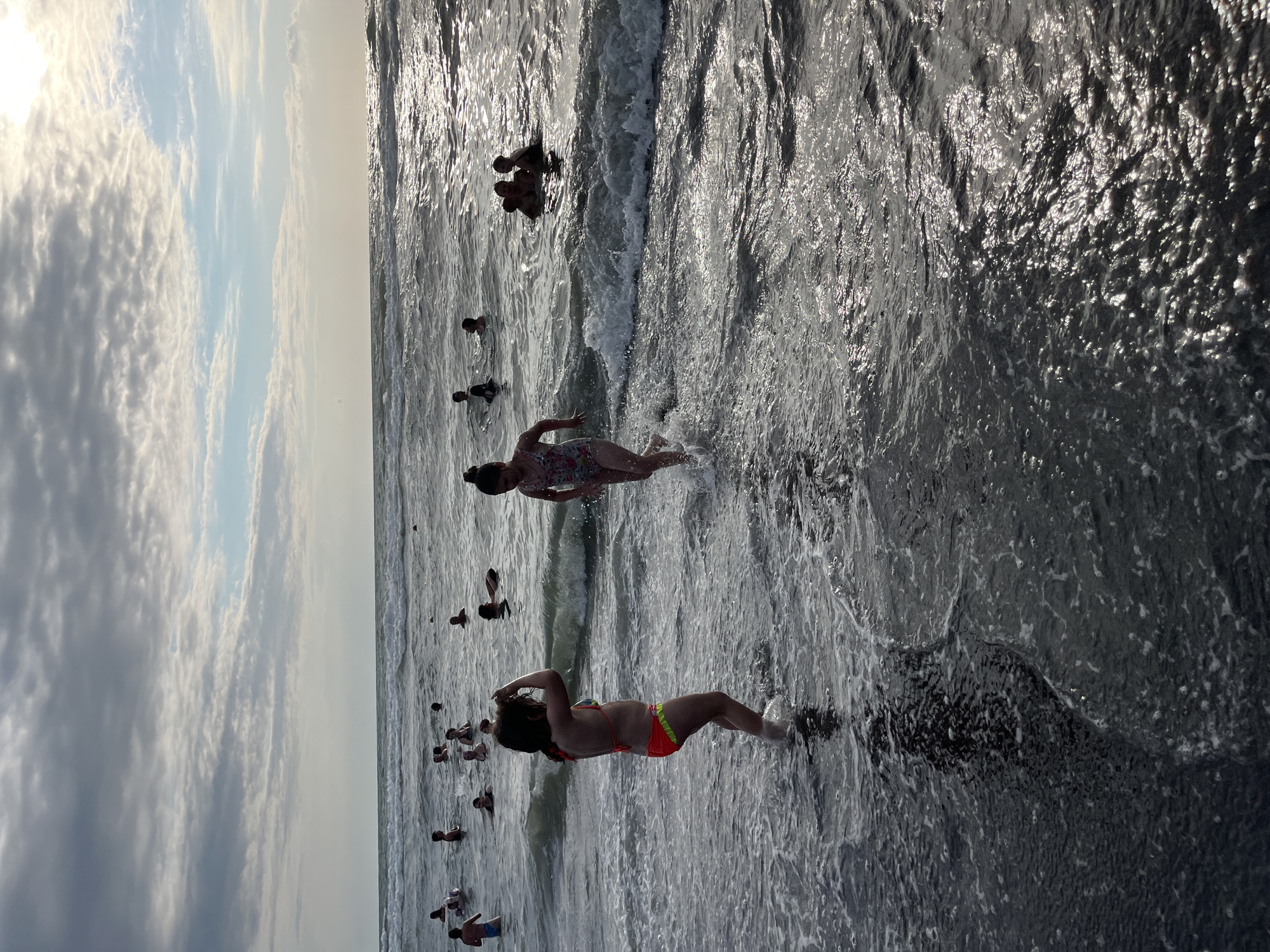 The image depicts a lively beach scene in Ureki, Georgia. Several people, including children and adults, are enjoying the water and the waves of the Black Sea. Some are wading in the shallows, while others are further out, swimming or playing in the surf. The sun is high in the sky, indicating it is likely midday, and the weather appears to be clear with some scattered clouds. The water is a shimmering gray, reflecting the sunlight, and appears to be relatively calm with small waves breaking near the shore. The overall atmosphere is one of relaxation and fun, with people embracing the warm weather and the opportunity to cool off in the sea. There are no visible objects or text in the image.