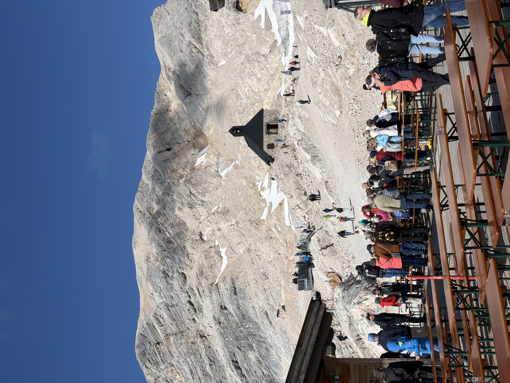 This image depicts the Zugspitze, Germany's highest peak, located near Garmisch-Partenkirchen. A group of tourists are gathered on a sun-drenched, rocky mountainside. In the mid-ground, a small, dark-roofed building with a steeple, possibly a chapel or a small restaurant, is visible. People are seen walking on paths and a viewing platform, some standing and observing the scenery, while others are seated at long, wooden picnic tables in the foreground, suggesting a dining or rest area. The mountain itself is imposing, with rugged, grey rock faces and patches of snow, indicating a high altitude. The sky is a clear, vibrant blue. The atmosphere appears to be lively, with many individuals enjoying the mountain environment.