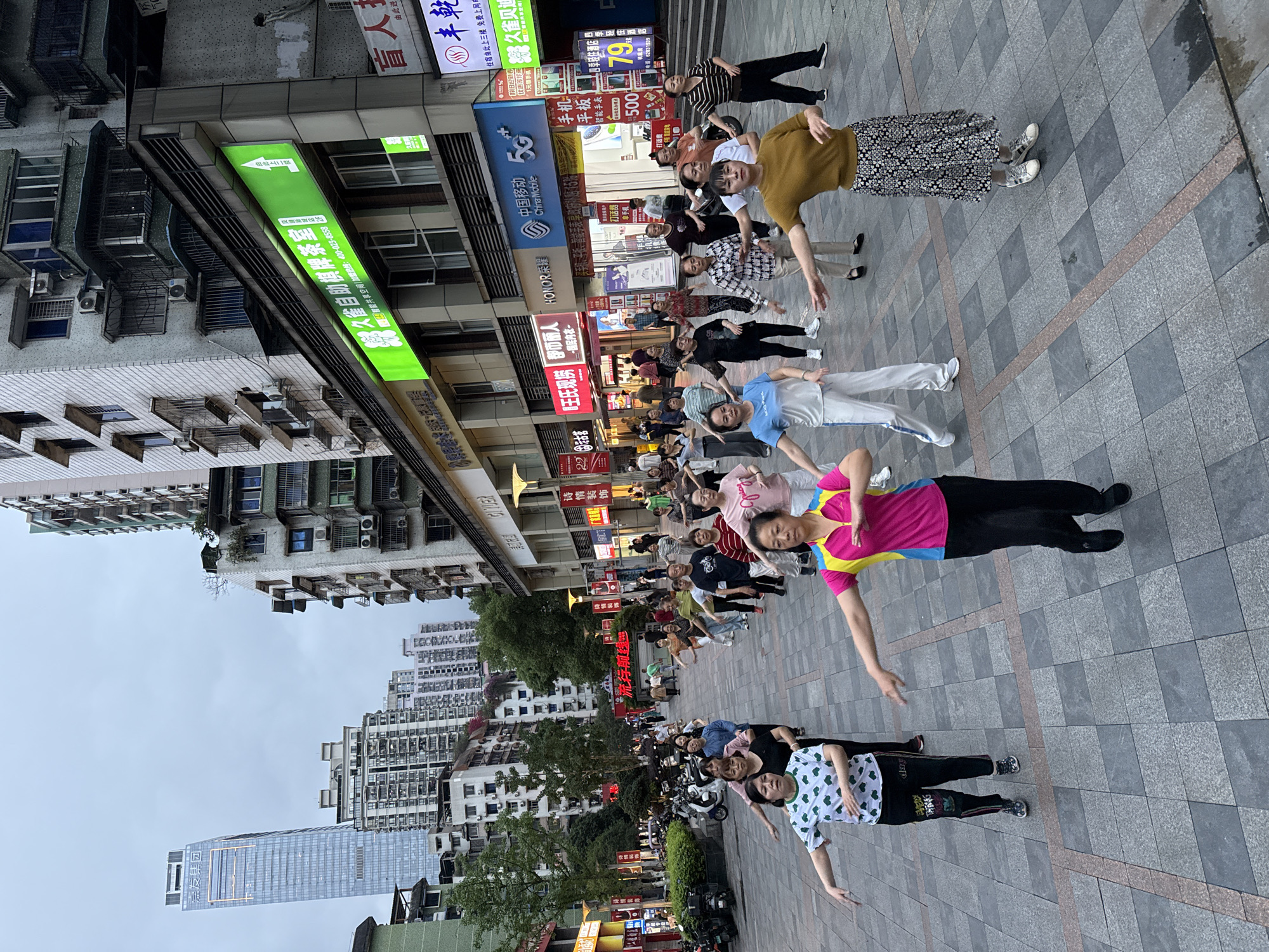 Here is a description of the image:
The image shows a group of people lined up in a row on a gray tiled pedestrian walkway, performing some sort of coordinated arm movements, possibly a dance routine or exercise. The participants appear to be mostly older adults. They are dressed in casual attire.
The scene appears to be an urban environment, likely a commercial area, with buildings visible on both sides of the walkway. The buildings have various commercial signage in Chinese characters. Banners and advertising are visible as well. A taller skyscraper is visible in the background.
The OCR text from the image includes references to brands and services available in China, such as China Mobile 5G. The text also mentions that a location is in Nan'an District, China.