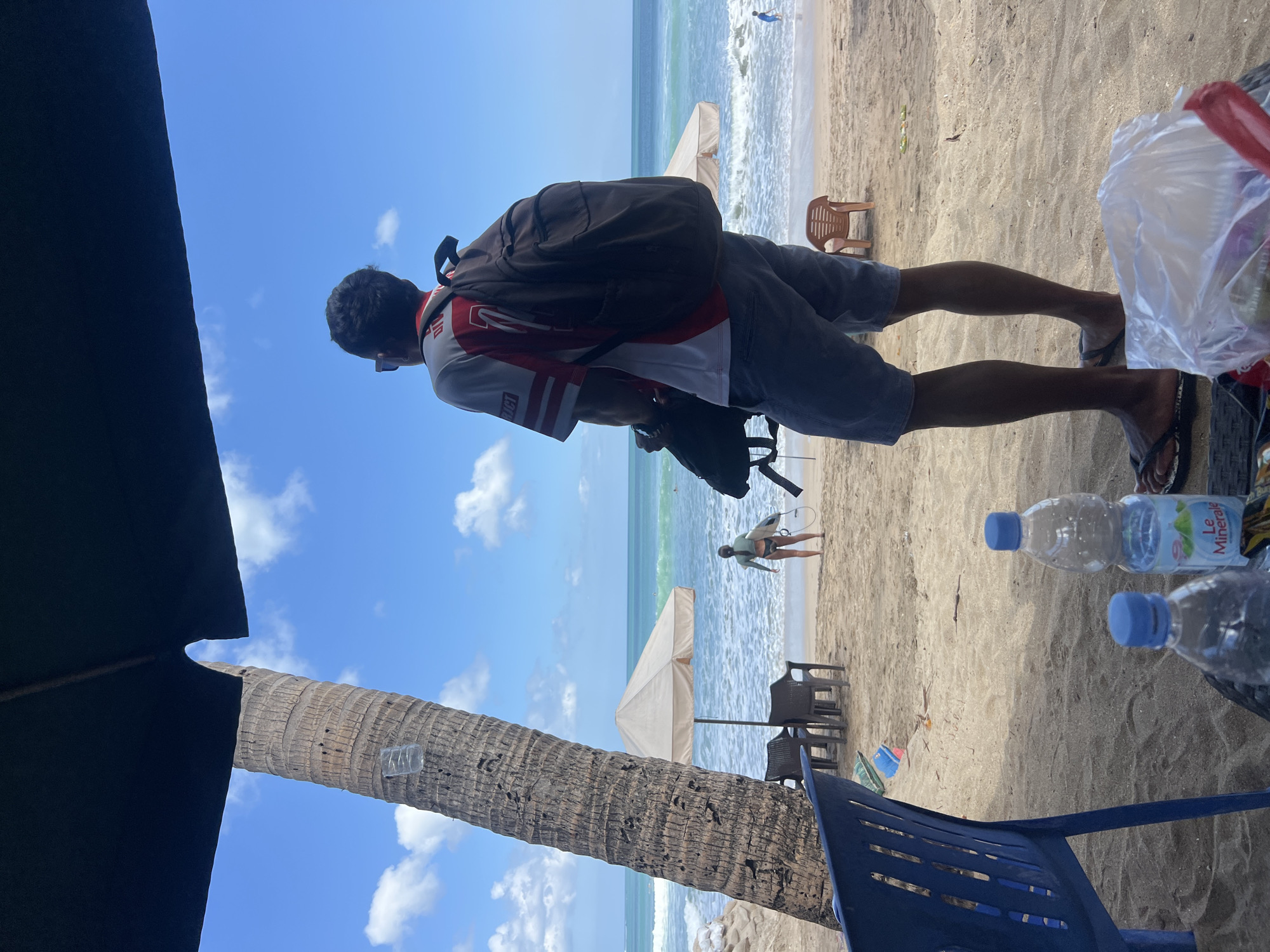 An eye-level, outdoor medium shot captures a man from the back, standing on a sandy beach in Kuta, Indonesia. He is wearing a white and red t-shirt, blue shorts, and flip-flops, with a dark backpack slung over his shoulders. To his right, a woman with a surfboard stands facing the ocean. In the background, the turquoise water of the sea meets a clear blue sky with scattered white clouds. The foreground is partially obscured by a dark umbrella, and several plastic chairs and water bottles are visible on the sand. The scene suggests a relaxed beach atmosphere with people enjoying the ocean and the sun.