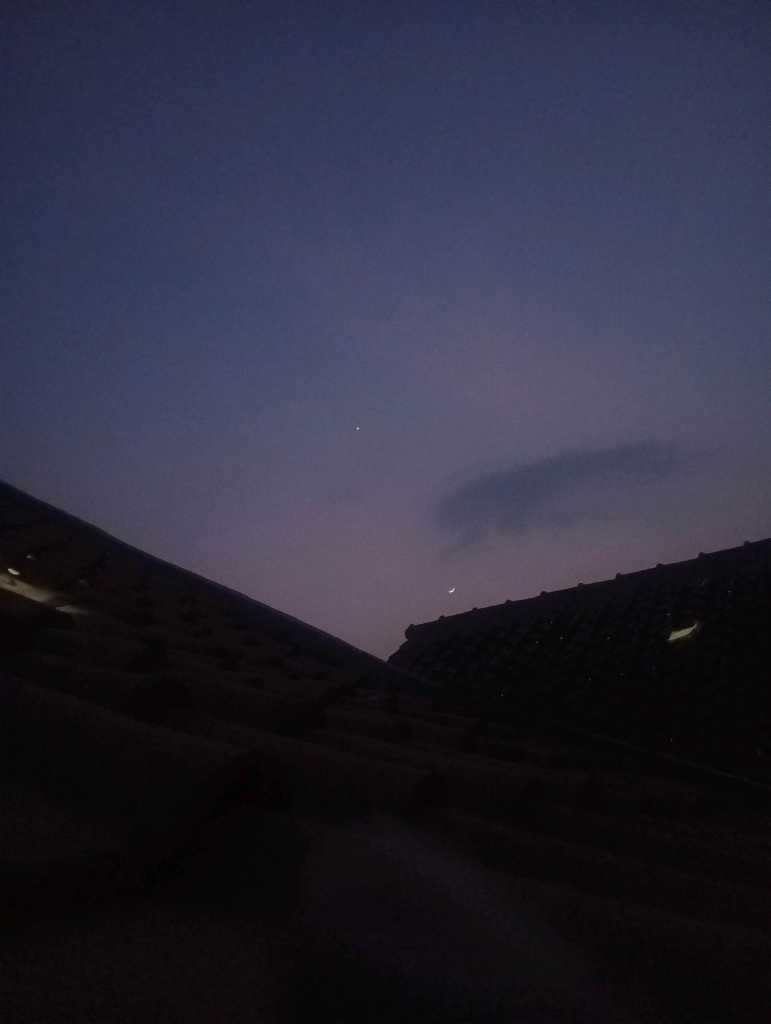 The image shows a twilight sky over rooftops in Kebandingan, Indonesia. The sky is a deep purple fading to a lighter hue near the horizon. A crescent moon is visible on the right side of the image, and a bright star or planet is visible higher in the sky on the left. The foreground consists of dark, silhouetted rooftops, appearing to be made of terracotta tiles. One rooftop on the left has a light source shining from beneath it, suggesting it is illuminated from within a building or from a light fixture. The overall atmosphere is calm and serene, capturing the transitional moment between day and night.