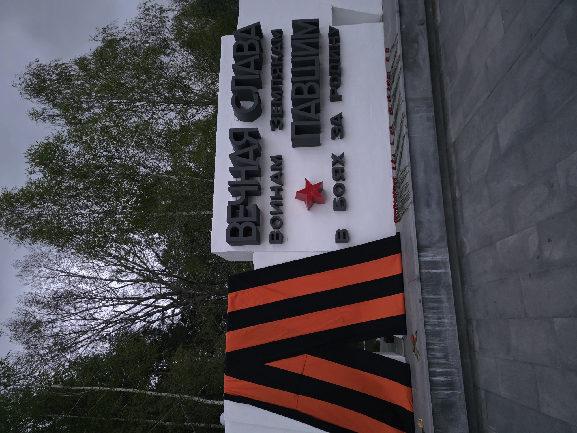 The image shows a war memorial in the city of Chaikovsky, Russia. The memorial is white with black lettering in Russian, featuring a red star. A black and orange striped ribbon adorns the base of the memorial. Red flowers have been laid at the base of the memorial. Trees are visible in the background under a cloudy sky. The image is rotated clockwise.