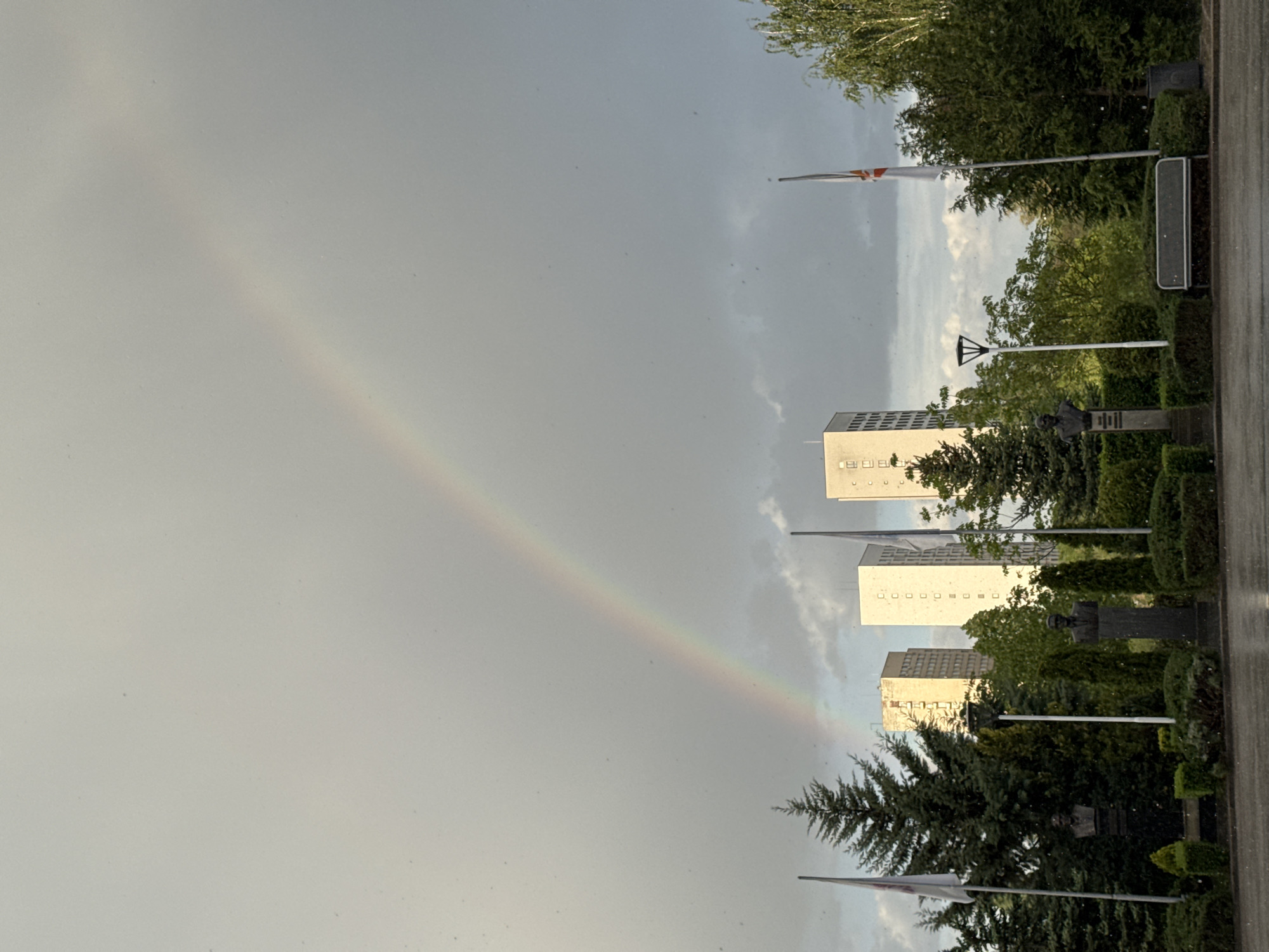 The image shows a segment of a rainbow in the sky. In the foreground, there are trees, flag poles and buildings. The buildings appear to be residential and likely of mid-rise construction.
The location is in Sofia, Bulgaria based on the building architecture.