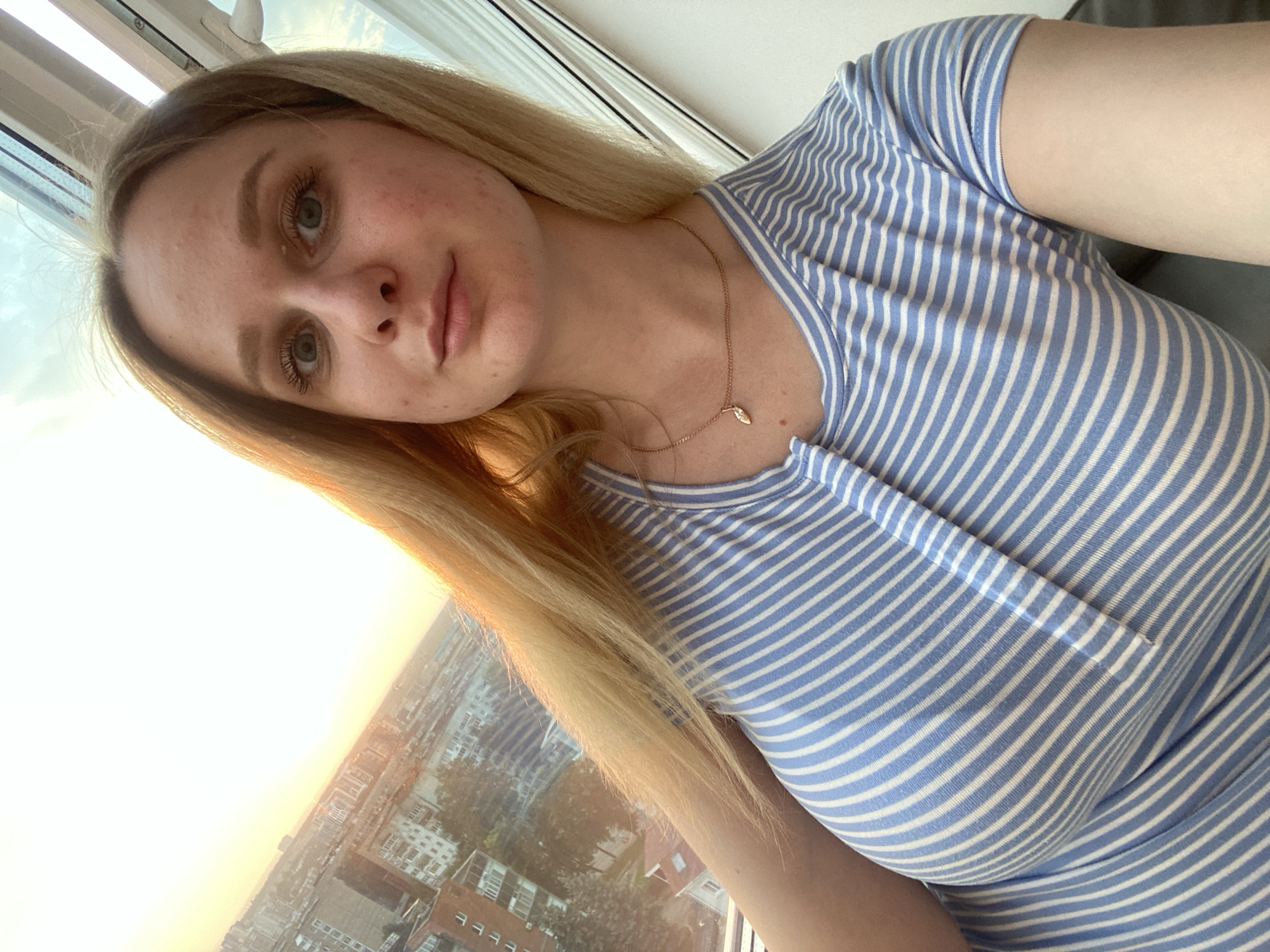 A young woman with blonde hair and blue eyes is captured in a selfie taken from a high vantage point. She is wearing a blue and white striped t-shirt and a delicate gold necklace with a small pendant. The background reveals a cityscape, likely Izhevsk, Russia, with various buildings and a hazy sky illuminated by what appears to be sunset or sunrise. The image seems to have been taken indoors, possibly from an apartment or hotel room, with a window providing the view.
