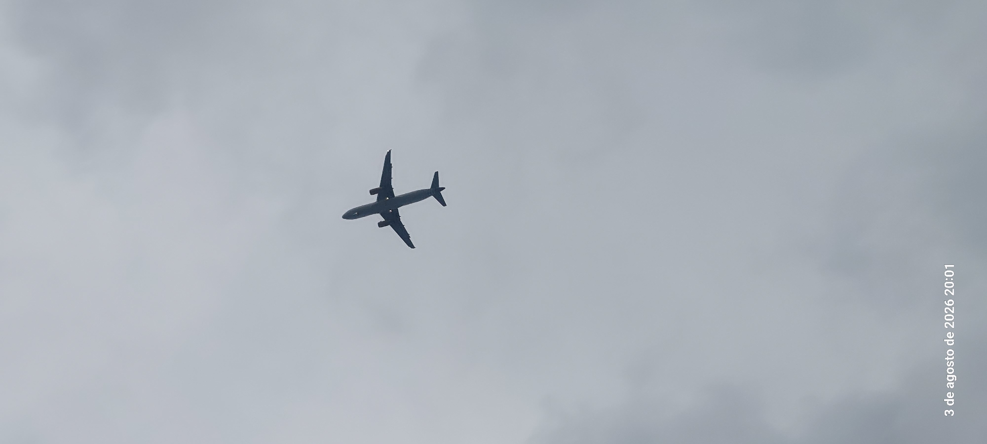A silhouette of an airplane is captured against a cloudy sky, appearing to fly from left to right. The image also displays a timestamp in the bottom left corner: "3 de agosto de 2026 20:01". This timestamp indicates the photograph was taken on August 3, 2026, at 8:01 PM. Based on the timestamp and the typical flight paths in the region, the photograph was likely taken in the vicinity of Castelldefels, Spain, as airplanes often fly over this area when approaching or departing from Barcelona–El Prat Airport.