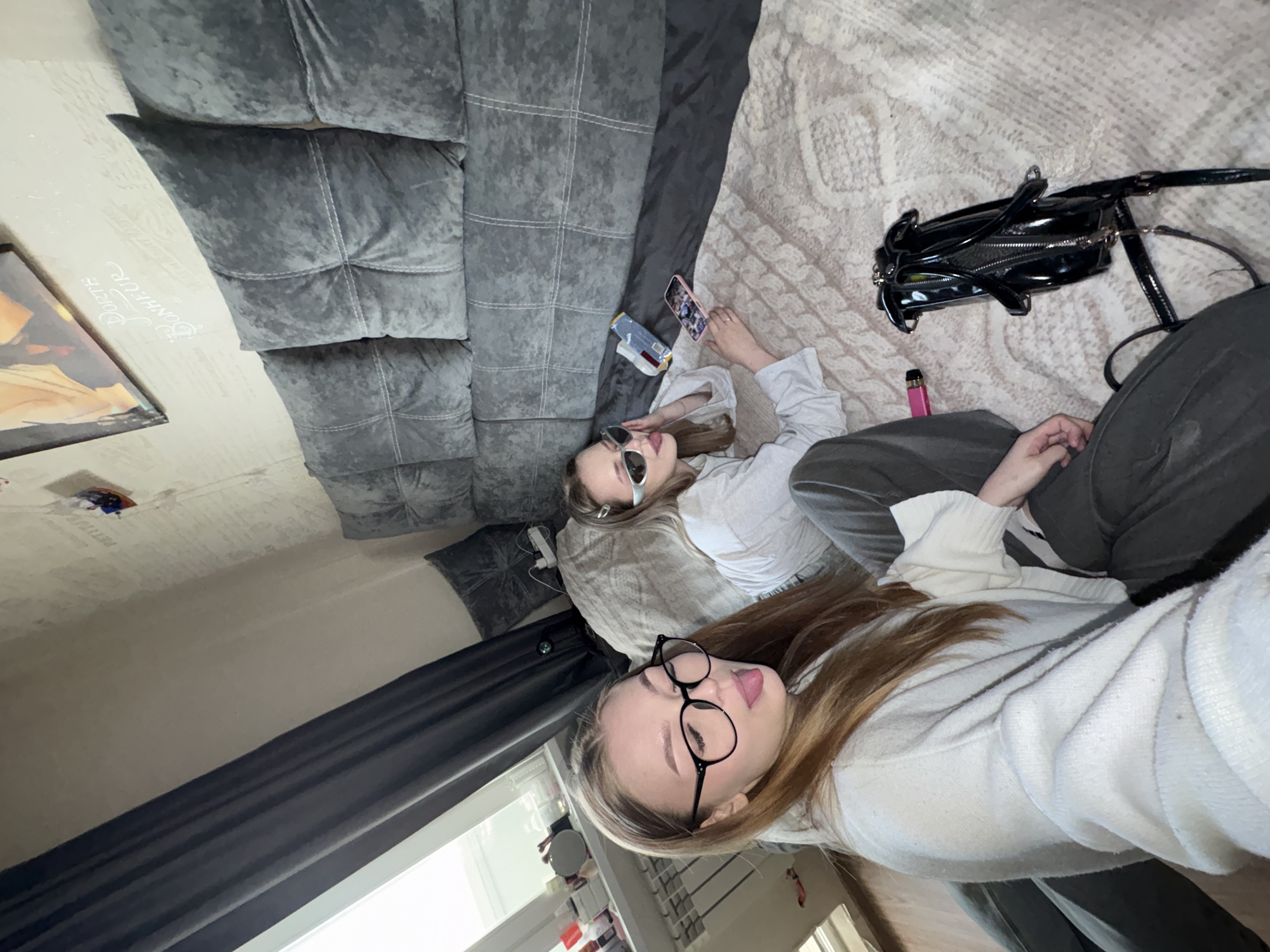 This high-angle, rotated image captures two young women relaxing indoors, likely in a living space or bedroom in Chaikovsky, Russia, during daytime hours. The photo is rotated 90 degrees clockwise.
In the foreground, a woman with long brown hair, wearing black round glasses and a light-colored knitted sweater, playfully sticks out her tongue towards the camera. She is dressed in dark trousers. Behind her, a second woman with long brown hair, wearing sunglasses and a white long-sleeved top with dark trousers, lies back in a more relaxed pose, looking away from the camera.
They are situated on a light-colored, patterned blanket or rug. To their left, a large, dark grey velvet sofa with prominent stitching provides a backdrop. Several personal items are visible: a black shiny handbag with a zipper and a pink vape pen are near the women's legs. A mobile phone with a pink case, displaying a gallery of images, and a small packet (possibly snacks) are placed next to the woman in sunglasses.
The wall in the background is light-colored, featuring cursive text that reads "Dolce Bonheur," a phrase meaning "sweet happiness." A partially visible framed picture hangs on the wall. Dark grey curtains cover a window, letting in natural light. The overall scene suggests a casual and relaxed atmosphere, capturing a moment of leisure between the two individuals.