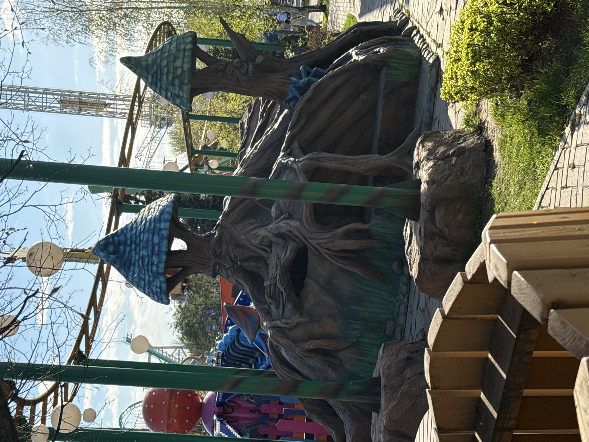 The image depicts an amusement park attraction in Khimki, Russia. The attraction appears to be a themed ride with large, artificial tree sculptures featuring peaked, blue-tiled roofs. A green metal structure, possibly part of the ride's framework, is visible in the foreground, along with decorative mushroom-shaped objects. In the background, a taller ride structure can be seen against a partly cloudy sky. Other park features, such as a walkway paved with rectangular stones and green vegetation, are also present. The scene is captured in daylight.