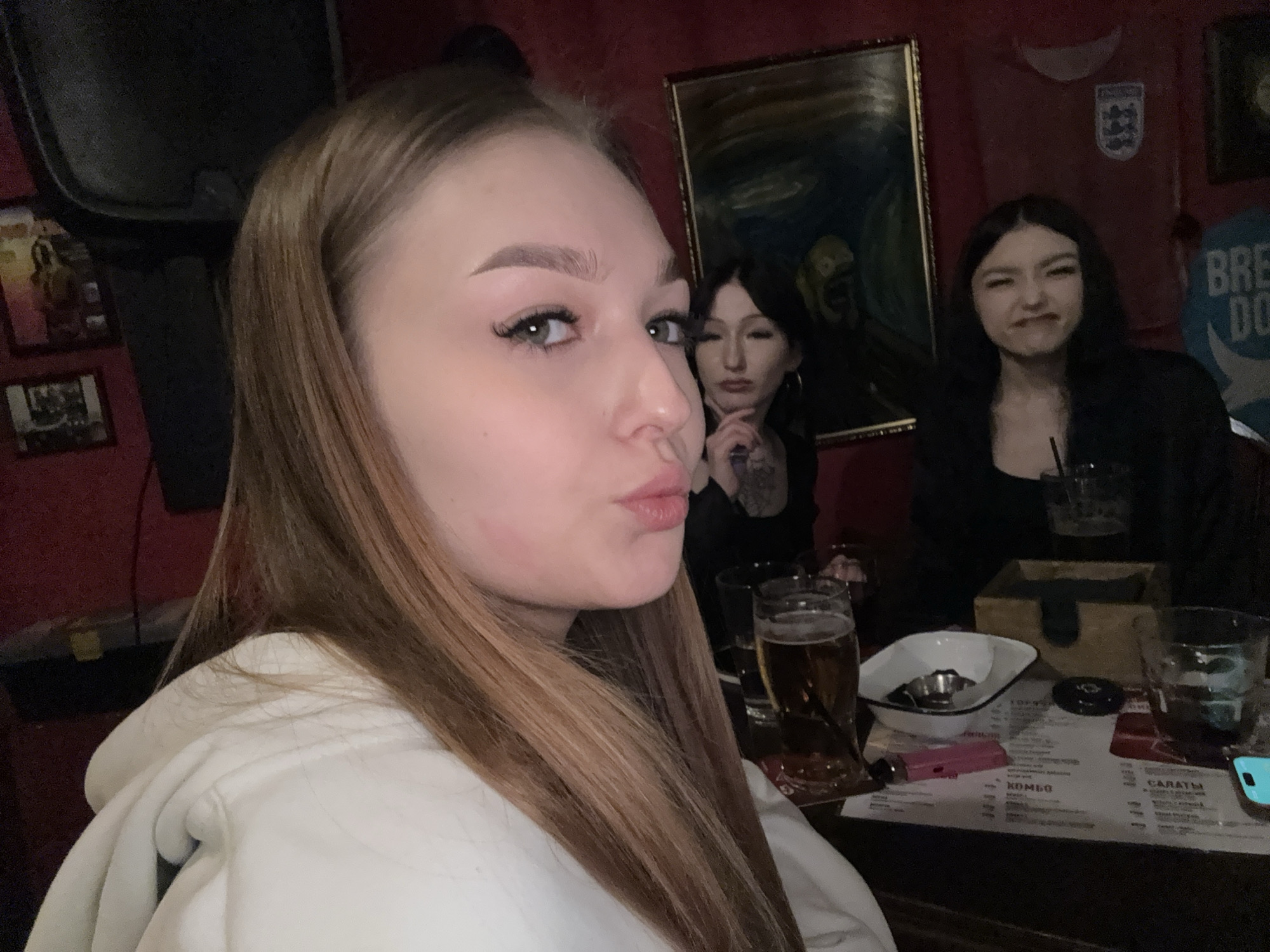 Three young women are at a table in what appears to be a bar or pub. The woman in the foreground has light brown hair and is wearing a white hoodie, making a kissing face at the camera. Behind her, another woman with dark hair and tattoos on her arm is looking slightly to the left with a serious expression. The third woman, with dark hair, is further back and making a slightly grumpy face.
On the table in front of them, there are drinks (likely beer), an ashtray, a lighter, and a menu. Behind them, the decor includes pictures on the wall, including a framed print reminiscent of Van Gogh's "The Scream," and an England soccer jersey. The environment suggests an informal setting, likely a social gathering. This image does not provide information to verify this location in the city of Chaikovsky, Russia.