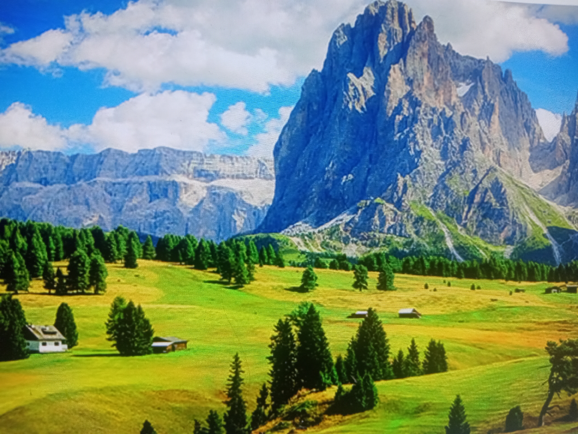 An eye-level, medium shot captures a vibrant landscape dominated by a majestic mountain range under a bright blue sky dotted with fluffy white clouds. The foreground transitions from rolling green hills, some with golden patches, to a lush green meadow. Scattered across the meadow are dark green evergreen trees, with a cluster of them in the middle ground to the right. Two small, rustic wooden buildings are visible in the middle ground, one to the left and another to the right. A white house with a dark roof is situated in the lower left quadrant, accompanied by a dark green tree to its right. The mountain peaks are rugged and textured, with some showing patches of snow.