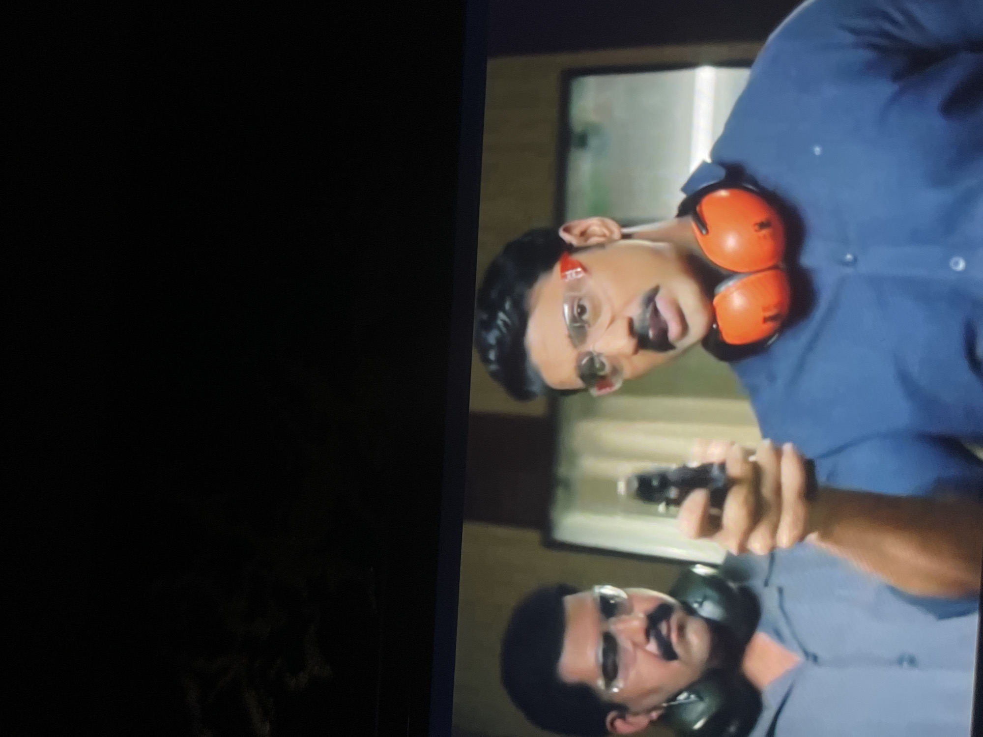 The image shows two men wearing safety glasses and earmuffs in what appears to be a shooting range. The man on the right is holding a handgun, aiming it forward. He is wearing a blue shirt and bright orange earmuffs around his neck. The man on the left, partially visible, is wearing a gray shirt and green earmuffs. The environment appears to be indoors with a plain background. There is no indication of the city of Bukuru, Nigeria.
