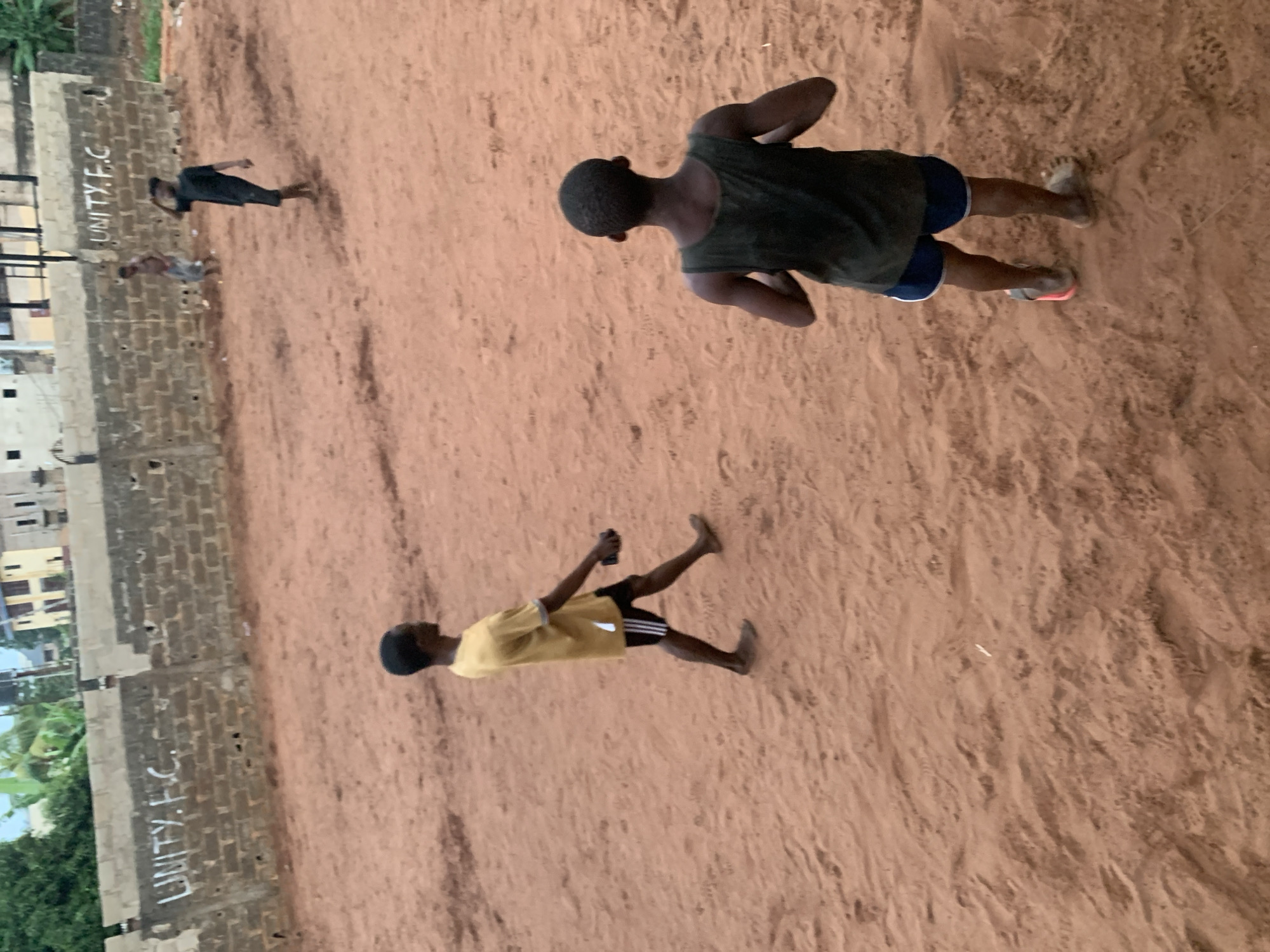 The scene is an outdoor reddish-brown sandy field. Along one side of the field is a gray brick wall with the words "UNITY F.C." written on it. Several individuals are present. One person, wearing a yellow shirt and black shorts with white stripes, appears to be walking or running. Another individual is wearing a dark tank top and dark blue shorts. Another person is wearing a dark shirt and dark shorts. The location is Ukwu-Abwa, Nigeria.