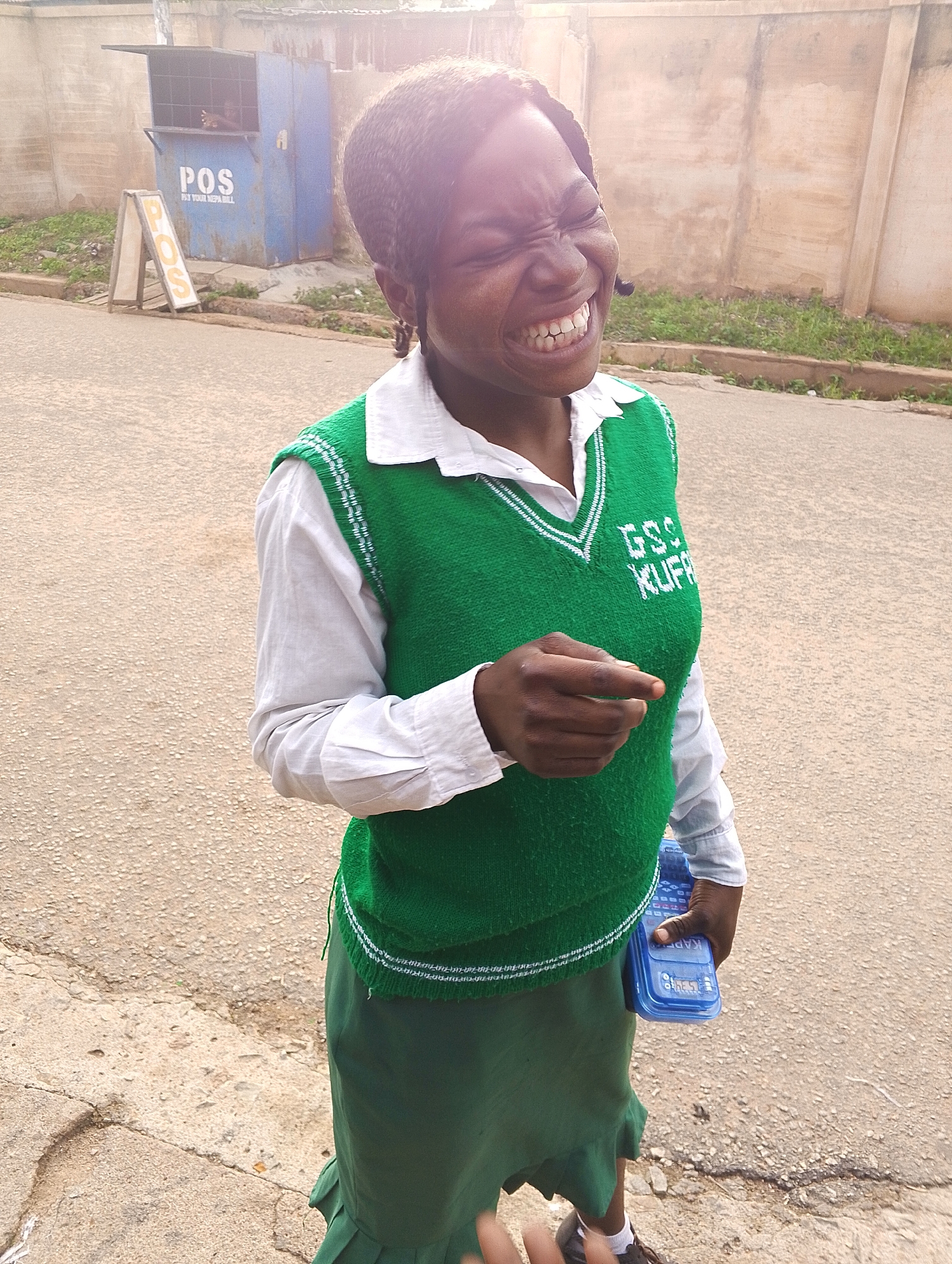 A young woman, with her eyes squeezed shut and a wide grin, stands on a paved surface, likely a street or road. She is wearing a white shirt and a green V-neck vest with white trim and the embroidered text "GSS KUFA". A dark green skirt reaches below her knees. She is holding a blue electronic device, possibly a calculator, in her right hand. In the background, a blue kiosk labeled "POS" with the words "PAY YOUR NEPA BILL" is visible, along with an A-frame sign that also says "POS". The scene appears to be outdoors, with a wall and some overgrown grass in the background. The lighting suggests it is daytime, with a bright sun creating a lens flare effect. The environment suggests a commercial area within Jos, Nigeria, given the "POS" and "NEPA BILL" signage, commonly found in such locations.