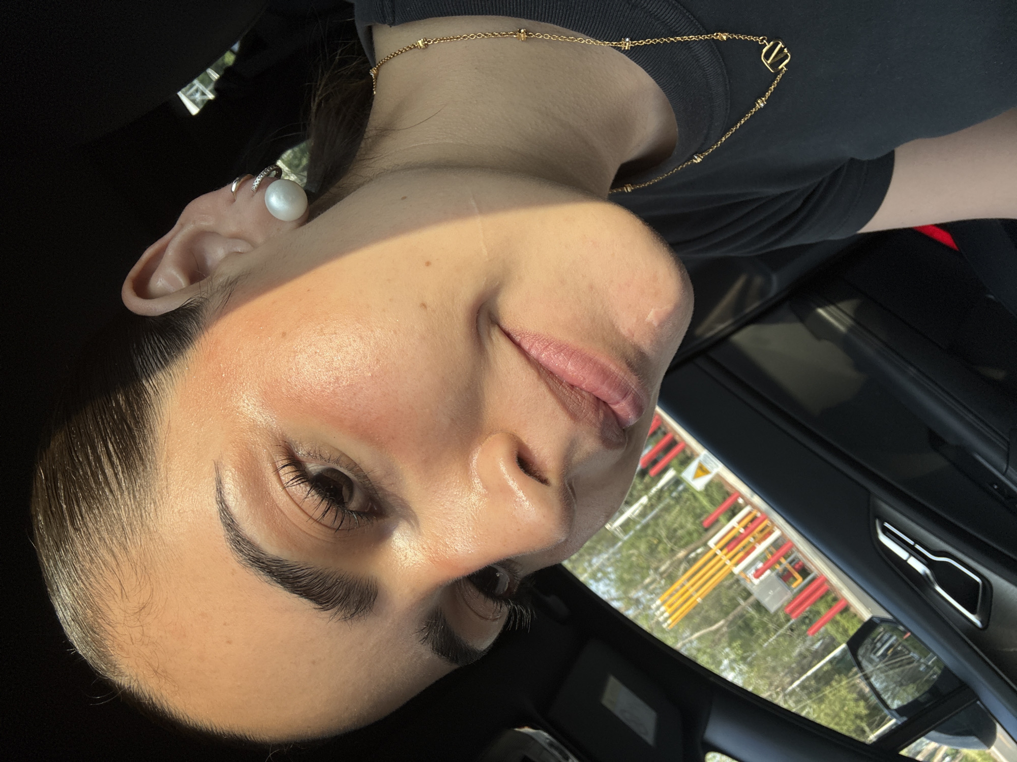 A close-up, rotated view of a woman's face is presented, seemingly taken inside a vehicle. She is wearing a black shirt and a delicate gold necklace featuring a "V" pendant. A large pearl earring is visible on her left ear. Outside the vehicle, through the sunroof, a street scene is partially visible, including vertical, colorful structures (red, white, and yellow). Based on the colors and structure of the seen elements, these structures appear to be a gas installation near the Atlixcayotl highway and Via Atlixcayotl in Puebla, Mexico. The interior of the car includes the passenger door and handle.