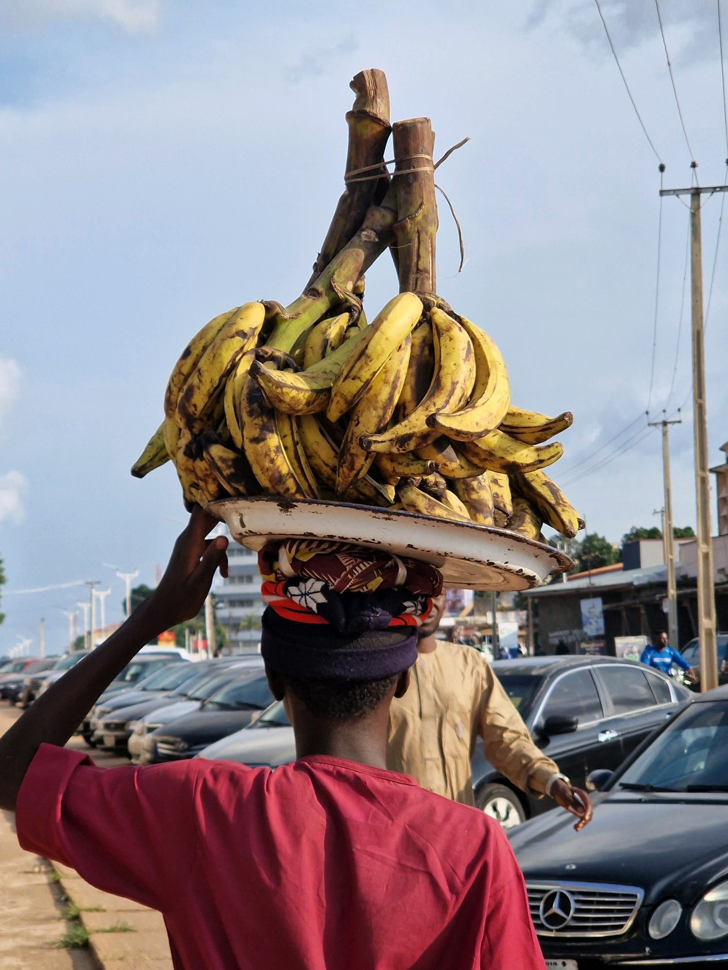 A person in a red shirt and dark headwrap walks with a large bunch of bananas balanced on their head. The bananas are attached to a wooden structure. The person's left hand reaches up to steady the bananas. In the background, several cars are parked along a street, and utility poles with wires are visible against a blue sky with clouds. Another person in a tan shirt stands behind the person with the bananas. The scene appears to be outdoors in an urban environment.