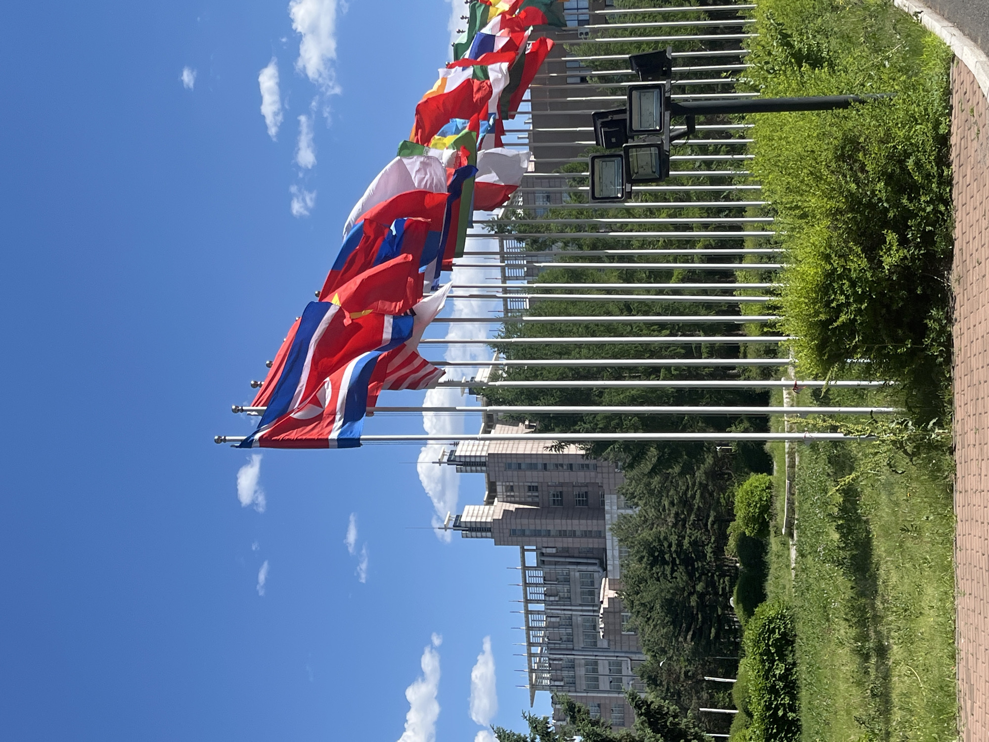 The image shows a row of international flags waving in the wind against a blue sky with scattered clouds. The flags are mounted on silver poles lined up along a grassy area. Behind the flags, there is a tall building complex with a modern architectural design. The scene appears to be outdoors on a sunny day. The flags are located in Changchun City, China.