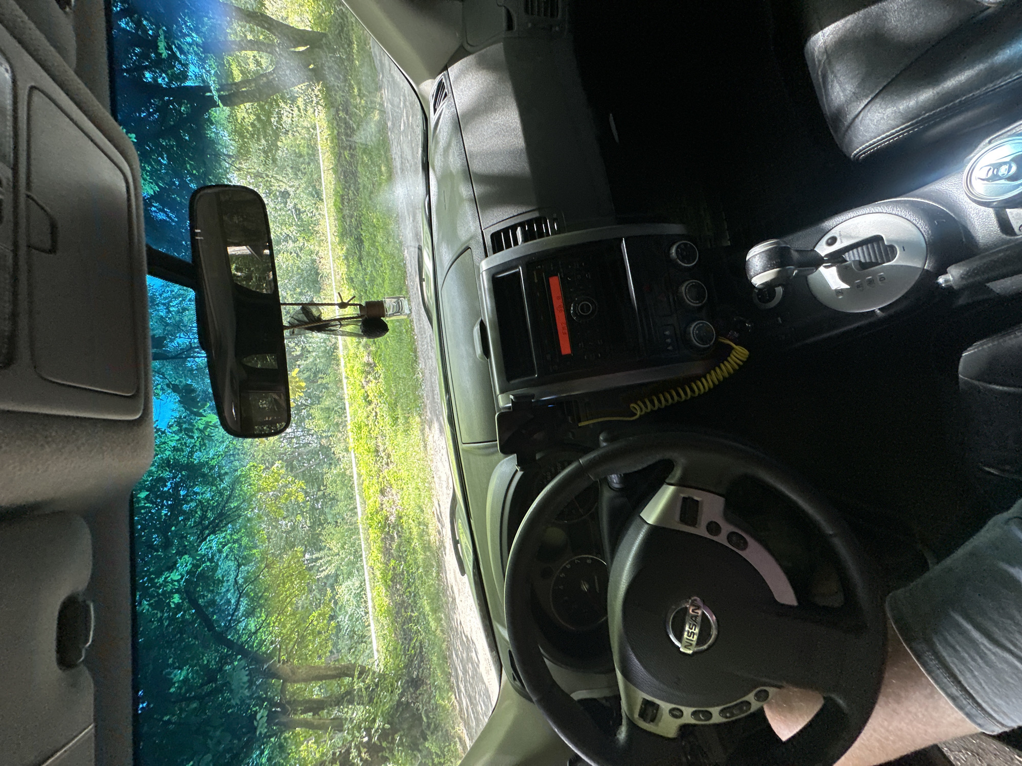 The image shows the interior of a car parked on a dirt road, likely in Shatava, Ukraine. The driver's side of the car is visible, including the steering wheel with a Nissan logo and various controls. The dashboard features a radio display showing "FM2 88.9" and a gear shift. Through the windshield, lush green trees and foliage are visible, suggesting a rural or park-like setting. The sun appears to be shining, casting dappled light on the interior and exterior. There is a hanging air freshener from the rearview mirror. The overall impression is of a vehicle at rest in a natural environment.