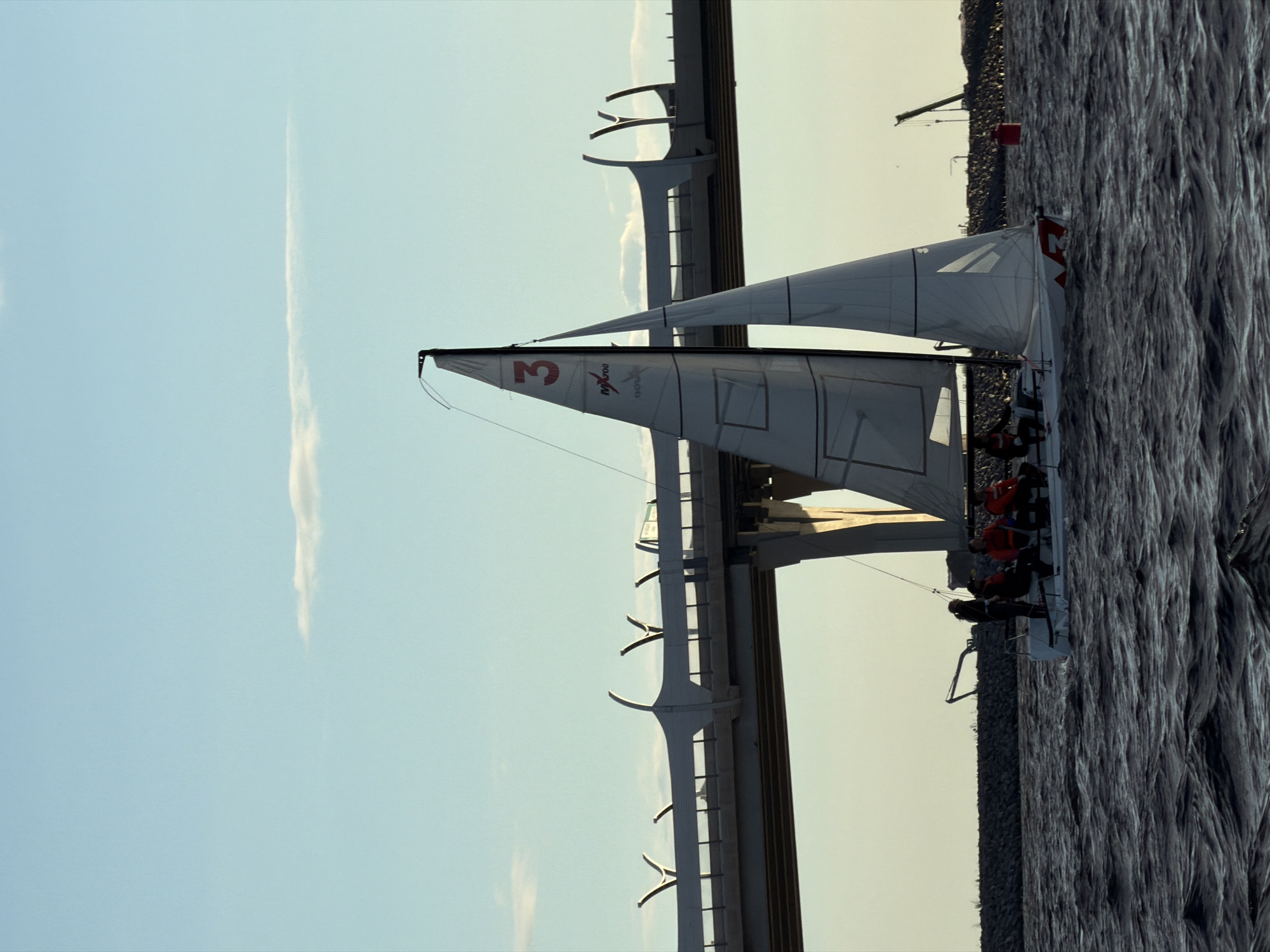 In Saint Petersburg, Russia, a sailboat navigates choppy dark water with a modern bridge and a segment of coastline in the background. The sailboat, white with red accents, displays the number "3" prominently on its sail. Four individuals are visible on the sailboat's deck, some wearing life vests. The bridge, a significant piece of infrastructure, features unique curved supports. The sky is a light blue with a few wispy clouds. The scene appears to capture a sailing event or recreational activity under a clear sky.