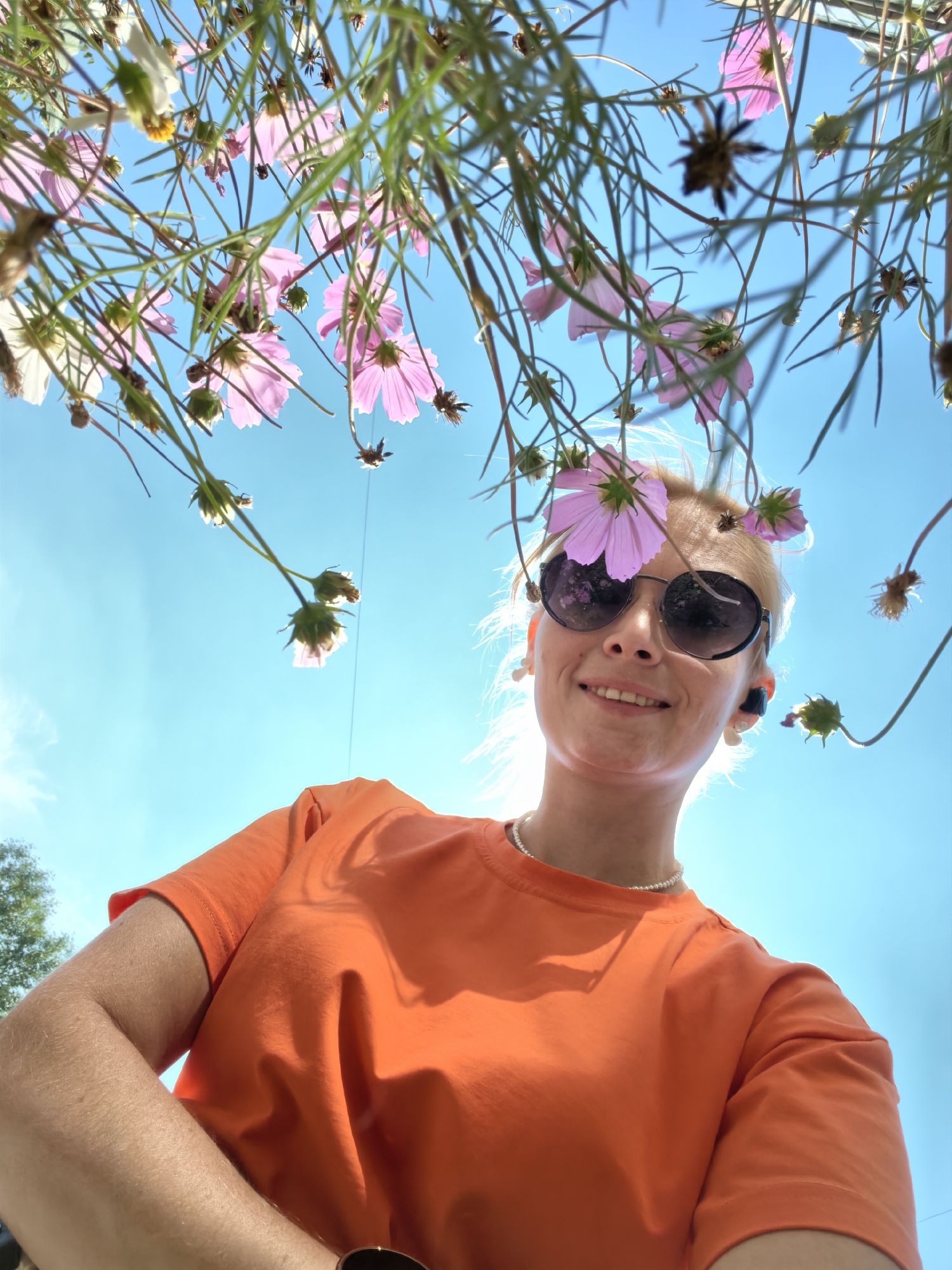 An outdoor, low-angle, close-up, selfie-style shot captures a smiling Caucasian woman with blonde hair wearing sunglasses and an orange t-shirt. She is positioned in the foreground, with pink and white flowers framing her head and the bright blue sky visible in the background. The image appears to have been taken on a sunny day, with the sun casting a glow on her hair. The location is specified as Sterlitamak, Russia.