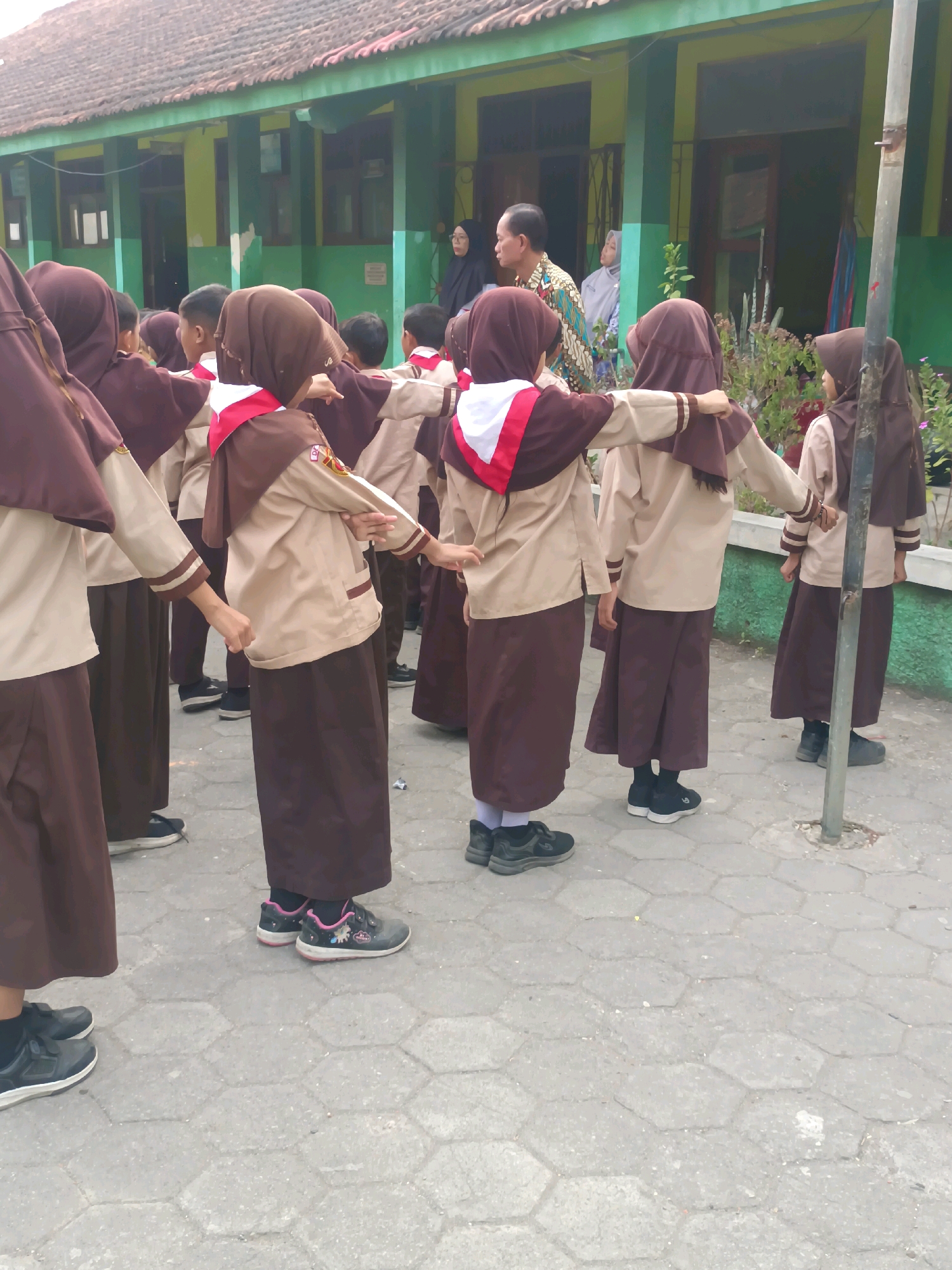In Sarimulyo, Indonesia, a group of schoolchildren in uniform are standing in formation outdoors. The students, predominantly female, are wearing brown skirts, tan shirts with brown trim, and brown headscarves. Some have red and white sashes across their chests. They are arranged in rows, facing away from the camera, and appear to be participating in an outdoor activity or assembly. In the background, a green school building with a tiled roof is visible. Adults, including a man in a patterned shirt and women in headscarves, are standing near the building. The ground is paved with hexagonal grey tiles. The overall scene suggests a school event or educational activity taking place in a tropical climate.