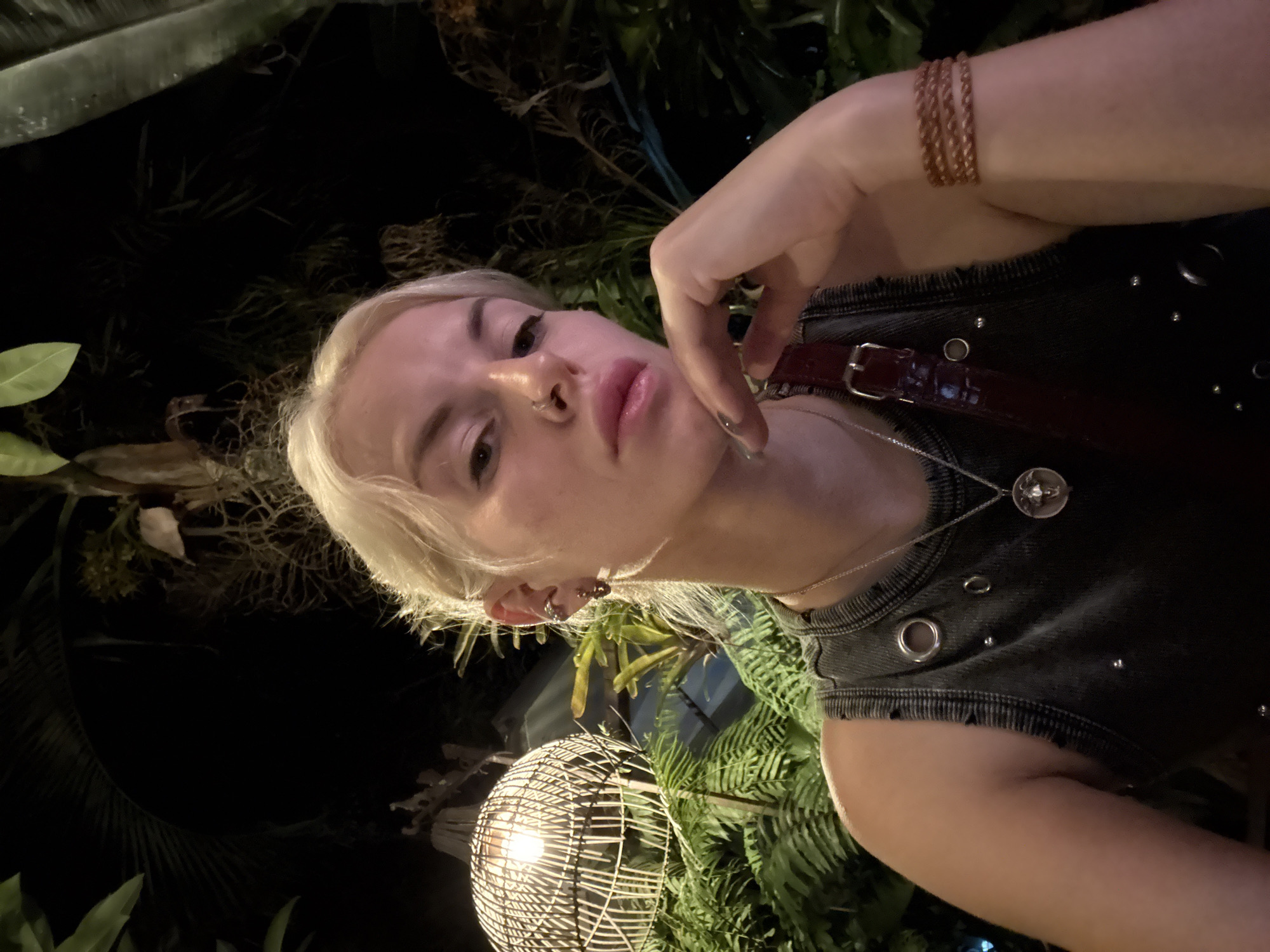An angled, close-up selfie captures a person with short, platinum blonde hair against a dark, lush tropical background at night in Baan Lamai, Thailand. The subject has light skin, a septum nose piercing, and wears a dark sleeveless top adorned with metal eyelets and studs. Their right hand, adorned with braided, copper-toned bracelets, is raised casually with fingers near their chin. A silver necklace with a circular pendant is visible, along with multiple earrings in the left ear.
The background features dense, dark green foliage, including ferns and broad-leafed plants, indicating an outdoor or garden setting. A prominent spherical, white woven or wicker lantern with a warm light source illuminates parts of the greenery and the lower portion of the frame. The overall lighting suggests a nighttime setting, highlighting the person and the decorative lantern amidst the dark, verdant environment. The person's expression is neutral and composed. No visible text is present.