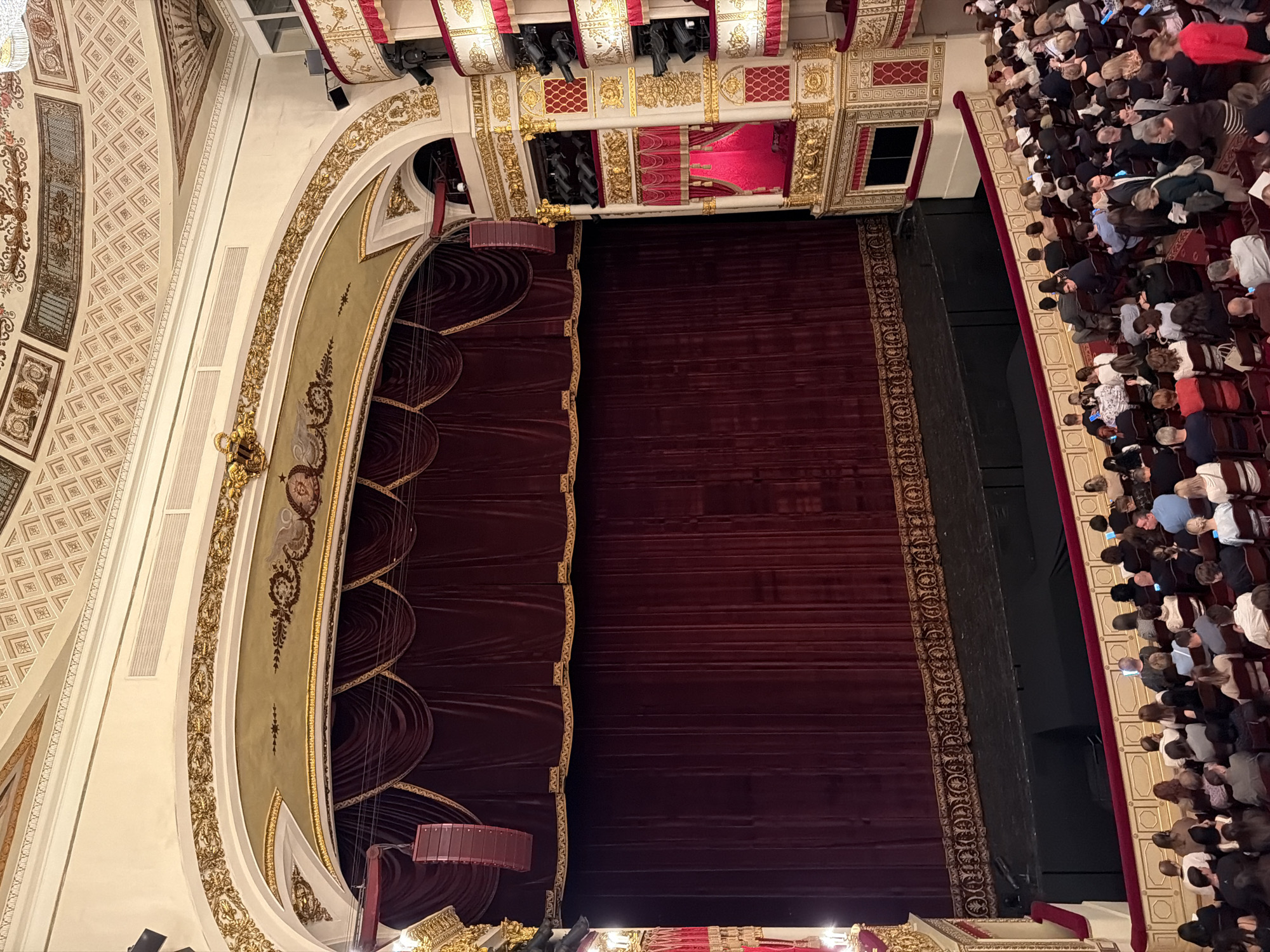 The image shows an interior view of a grand theater, likely the Mariinsky Theatre in Saint Petersburg, Russia, due to its opulent golden ornamentation, deep red velvet curtains, and tiered seating. The perspective is from a high vantage point in the audience seating area, looking towards the stage.
The stage is draped with heavy, dark red velvet curtains adorned with gold trim. Above the main curtain, a pelmet of the same material with tasseled drapery is visible. Two large, modern red loudspeakers are suspended from the ceiling on either side of the stage, indicating a contemporary use of the historic venue.
The auditorium itself features multiple levels of boxes and balconies, richly decorated with intricate gold detailing, white plasterwork, and red accents. The ceilings are ornately painted with geometric patterns.
In the foreground and extending across the bottom of the image, rows of theatergoers are seated. The audience members are dressed in a variety of casual to semi-formal attire. Many are looking towards the stage or at their mobile phones. The seating is arranged in curved rows facing the stage. The overall atmosphere suggests an event is about to begin or is in progress.