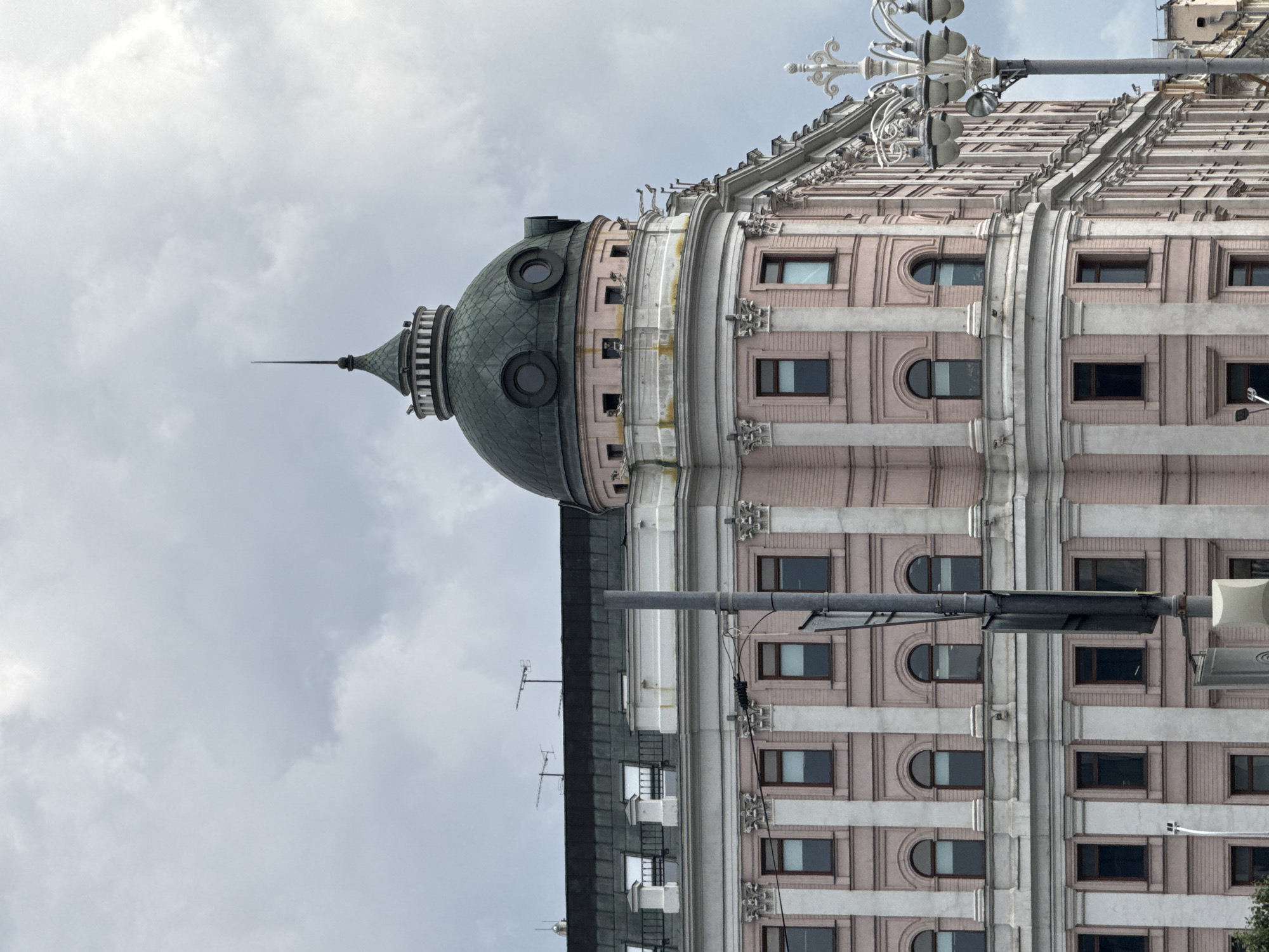The image shows a close-up view of a large, historic pink building with a green domed roof and a spire, located in Moscow, Russia. The building features classical architectural elements, including columns, ornate trim, and numerous windows, some of which are arched. The sky above is overcast with heavy clouds. A street lamp with multiple lights is visible on the right side of the frame, and a utility pole stands in front of the building. There are no people visible in the image. The overall impression is of an urban street scene with a grand architectural structure.