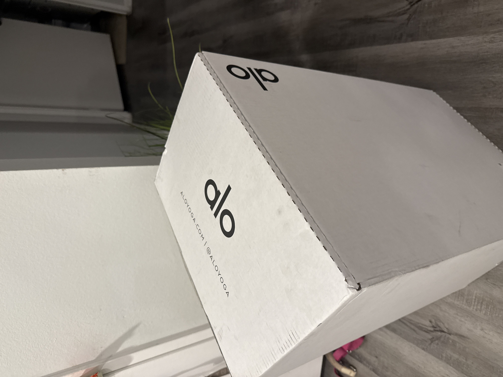A white Alo yoga package box is positioned on a gray wood-look floor next to a white cabinet. The box's side displays the brand's logo and website address. A blade of grass is visible near the box. The location appears to be indoors. The setting suggests a domestic environment, potentially in Los Angeles, USA, given the brand Alo Yoga's origin.