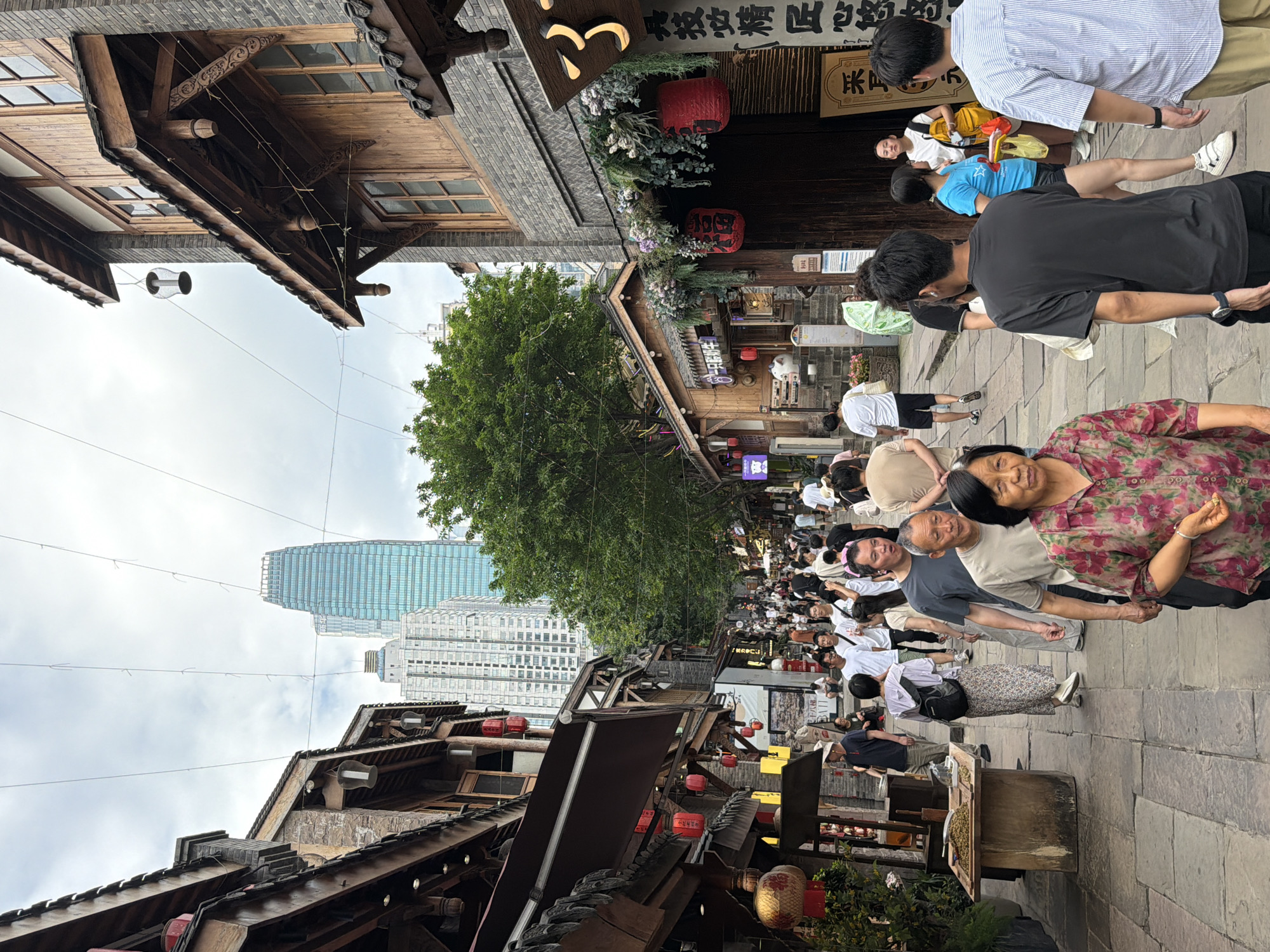 This image depicts a crowded street scene in Yuzhong District, China, likely during daylight hours. The street is lined with traditional-style architecture, characterized by wooden facades, tiled roofs, and red lanterns. In the background, modern skyscrapers pierce the overcast sky, creating a juxtaposition of old and new.
Numerous people are walking along the stone-paved street, some in small groups and others individually. They are dressed in casual, contemporary clothing. Several individuals are looking at their phones, while others appear to be engaged in conversation or browsing the shops lining the street. One person in the foreground is looking directly at the camera, their face visible.
Objects visible in the scene include market stalls with displays of goods, wooden tables with items on them, and various signage in Chinese characters. The overall atmosphere appears to be one of bustling commerce and pedestrian activity. The environment is a narrow, busy street with a sense of density and urban life.
