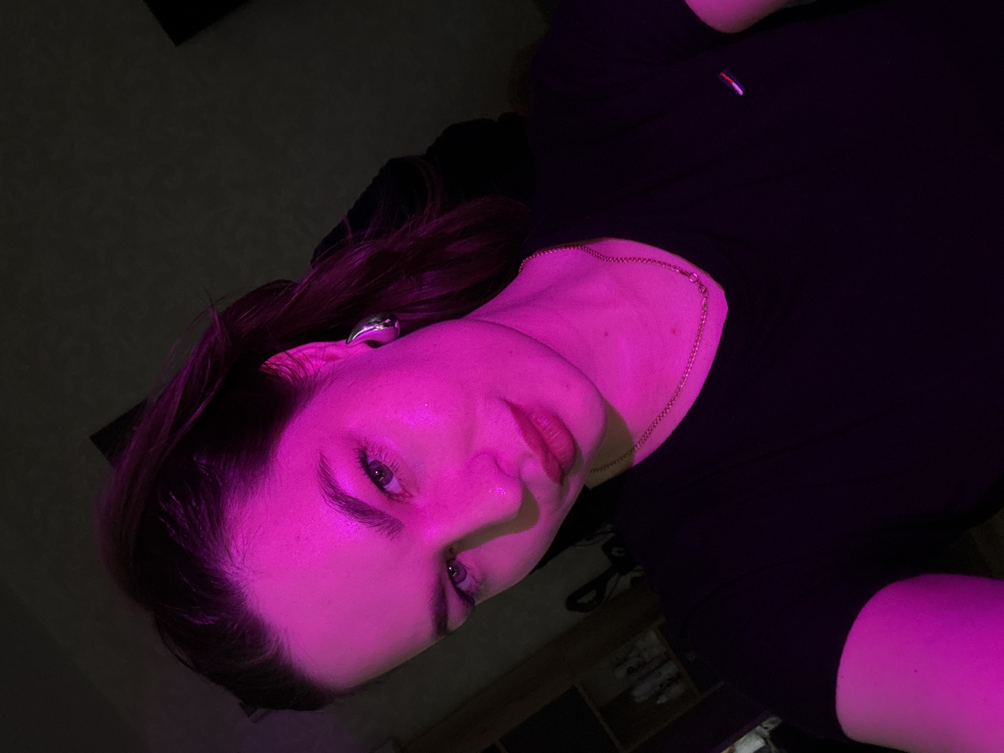 A medium close-up selfie of a woman is captured in a dimly lit room, illuminated by vibrant pink lighting. The woman, with her hair pulled back into a ponytail, is looking towards the camera with a neutral expression. She is wearing a black t-shirt with a small emblem on the left chest and a delicate gold chain necklace. A large, metallic earring adorns her left ear. The background is dark, with hints of shelves and bottles visible on the left side, suggesting an indoor setting, possibly a bedroom or dressing area. The pink light casts a strong, colorful glow on her face and shoulder, creating dramatic shadows and highlighting her features. The exact location within Chaikovsky, Russia, cannot be determined from the visual information provided in the image.
