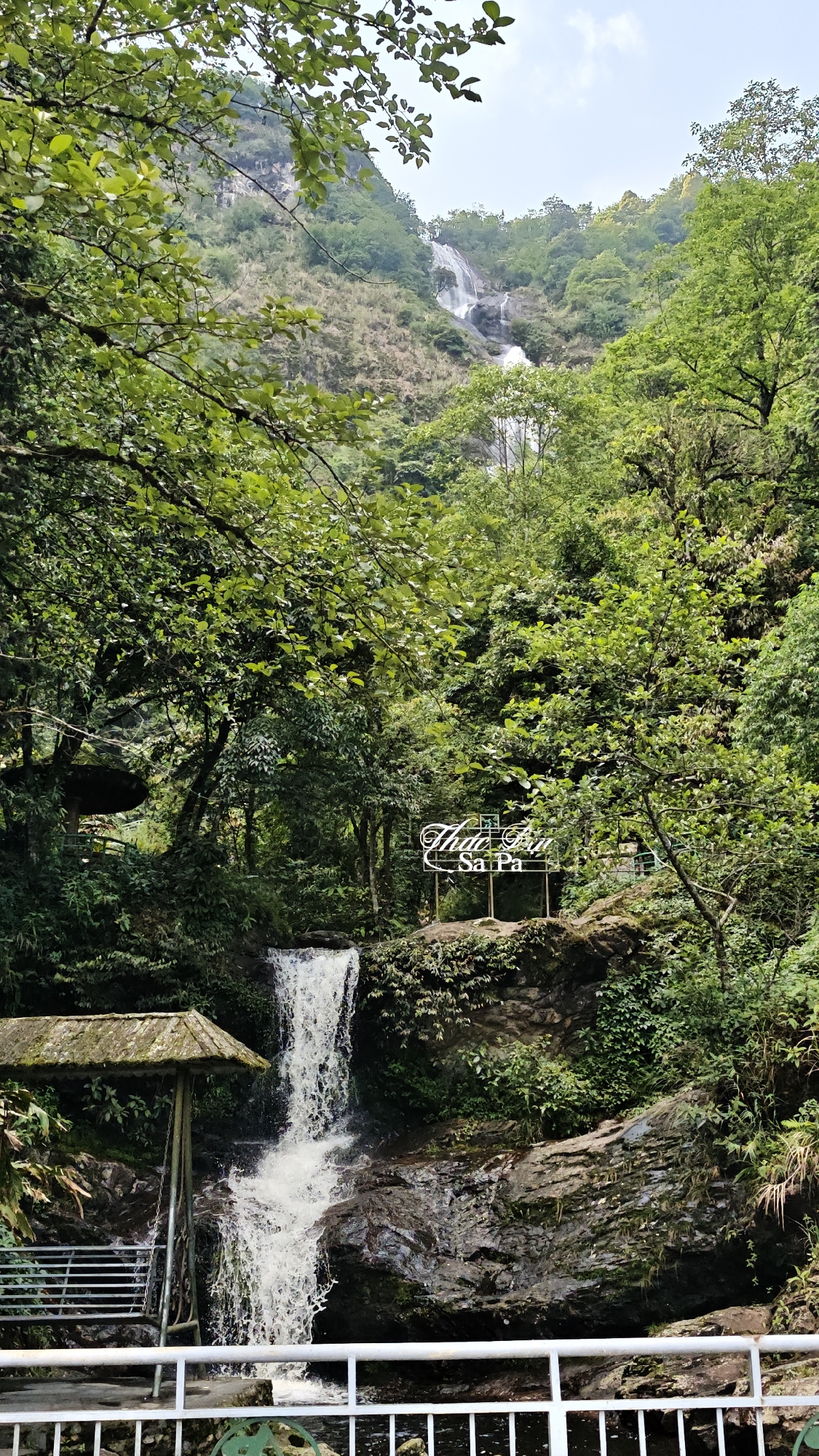 The image shows a landscape scene featuring a waterfall in Sa Pa Ward, Vietnam. The primary focus is on a lower waterfall cascading over rocks. A secondary waterfall is visible further up the slope in the background. Lush green vegetation and trees dominate the scenery. A structure with a thatched roof and seating is present in the foreground near the lower waterfall. A metal sign displaying "Thác Bạc Sa Pa" is visible. A white metal railing runs across the bottom of the frame.