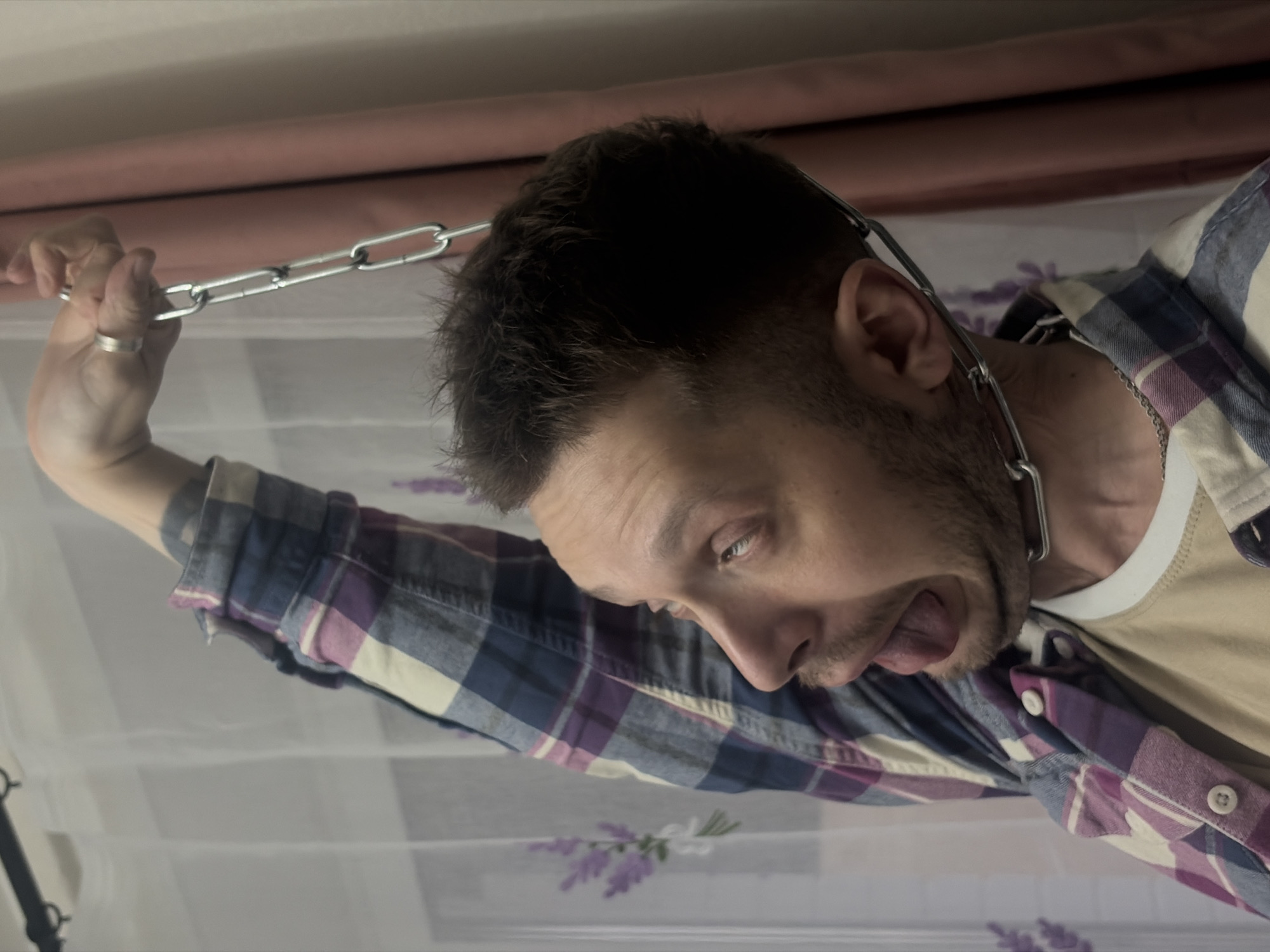 The image shows a man with short brown hair and a beard, wearing a white t-shirt and a blue and purple plaid shirt. He is holding a silver chain with one end near his neck and the other in his raised left hand. The man is looking down with his tongue partially sticking out. The background includes a peach-colored surface, a sheer white curtain with a floral pattern, and potentially a part of a camera stand. The location is not discernible from the image.