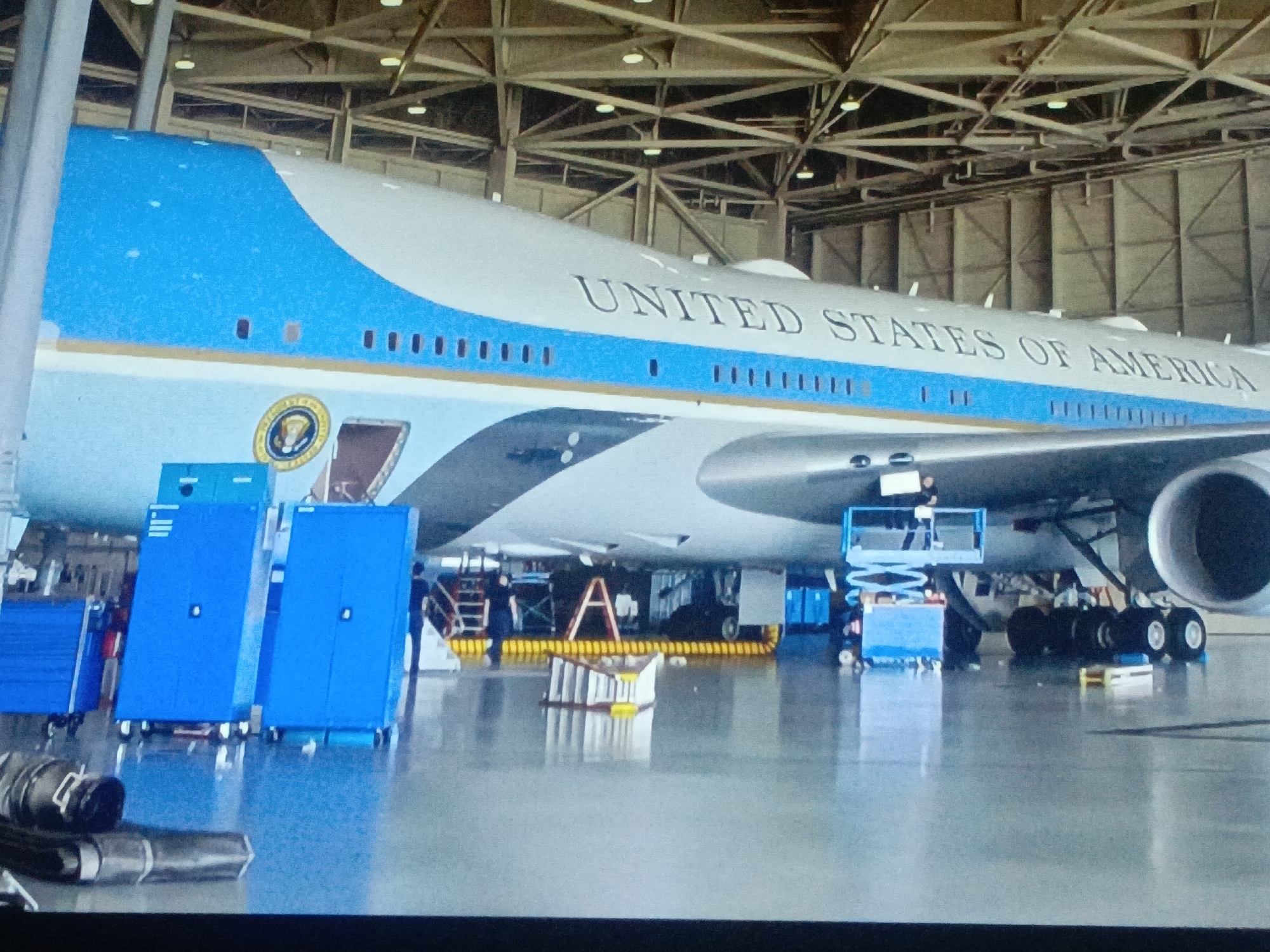 The image depicts Air Force One inside a hangar. Maintenance is being performed with various pieces of equipment including personnel lifts, toolboxes, and an A-frame ladder. "UNITED STATES OF AMERICA" is written on the side of the plane. The hangar has a high, crisscrossed beam ceiling. There is no indication of the location being in Juan Griego, Venezuela.
