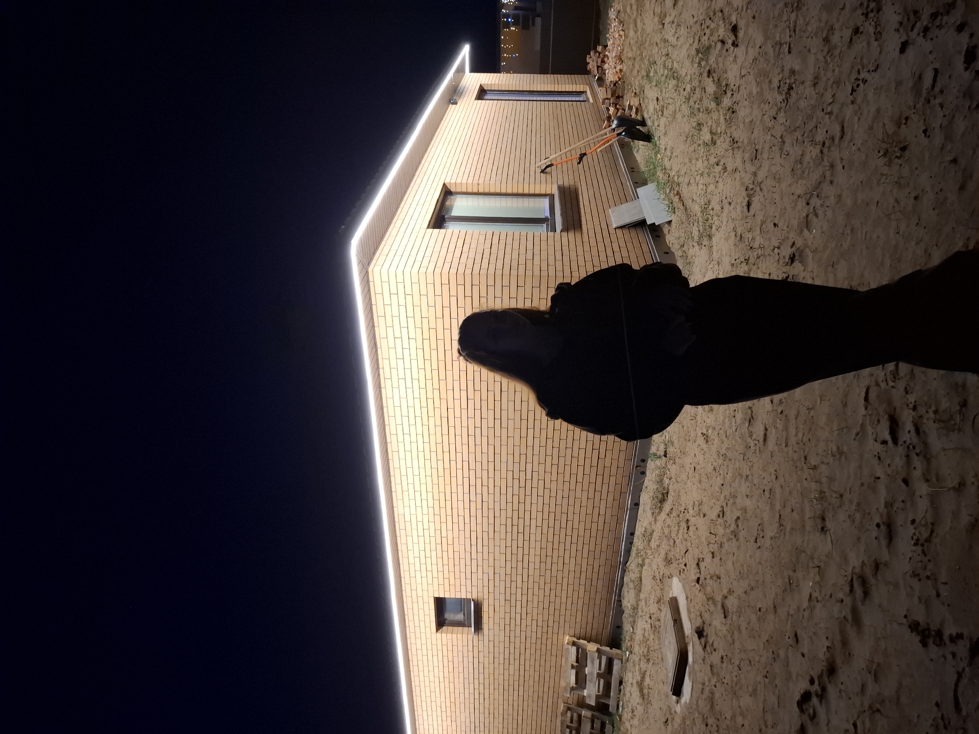 The image shows a brick building with white LED strip lighting along the roofline, set against a dark sky. The building has several windows. The ground appears to be sandy or dirt-covered, and there is a dark silhouette of a person in the foreground. There are also some wooden pallets and gardening tools visible near the building. The location is Chaikovsky, Russia.