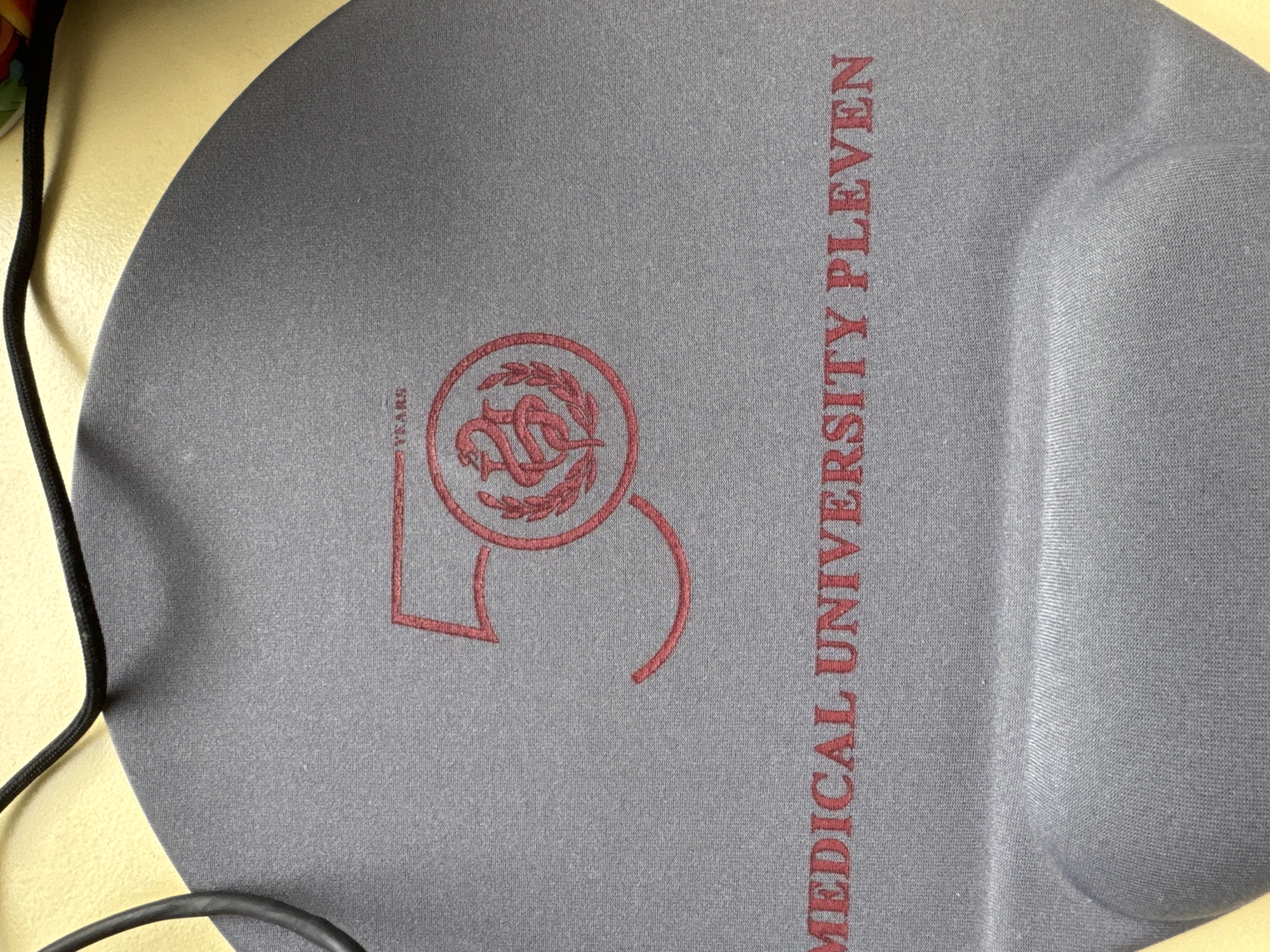 The image shows a close-up view of a dark gray mousepad. Embroidered on the mousepad in red are the number "50" with "YEARS" above it, and a circular emblem containing a caduceus. Below this is text that reads "MEDICAL UNIVERSITY PLEVEN". The mousepad has a wrist rest area at the bottom. The background is a yellow surface and a black cord is visible in the upper left corner. The image does not contain any information about the city of Smolyan, Bulgaria.