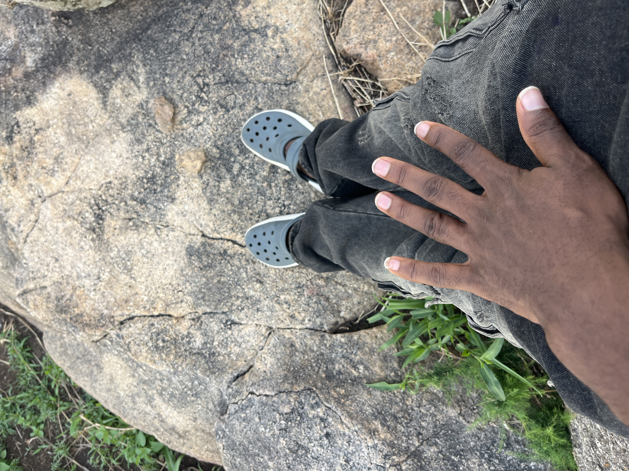 A person stands on a large, mottled grey rock with multiple visible cracks. The person wears dark grey Crocs and dark grey jeans. Their right hand rests on their right thigh. Green grass is visible at the edge of the rock and around the person's leg. The location is Jos, Nigeria.
