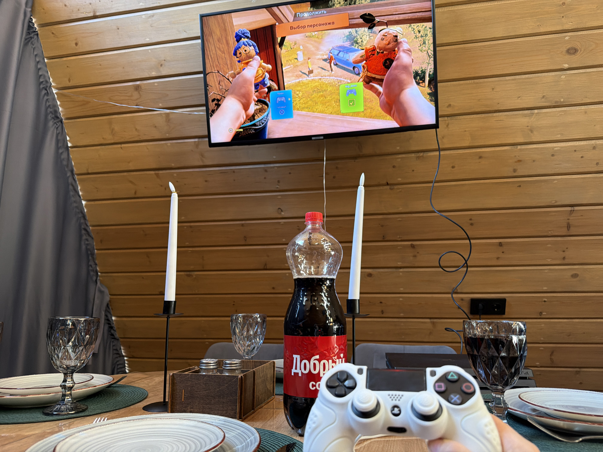 The scene appears to be inside a wooden cabin, possibly in or near Nizhnevartovsk, Russia, judging by the branding on the bottle. The cabin has wooden plank walls, and a dark grey curtain is visible on the left side of the frame. A flat-screen TV is mounted on the wall, displaying a video game where players select characters.
A dining table is set in the foreground with plates, silverware, and decorative goblets. A bottle of Russian "Dobryy Cola" sits between two tall, thin candles in black candle holders. A Playstation 4 controller is being held by a person in the immediate foreground, obscuring some of the table setting.