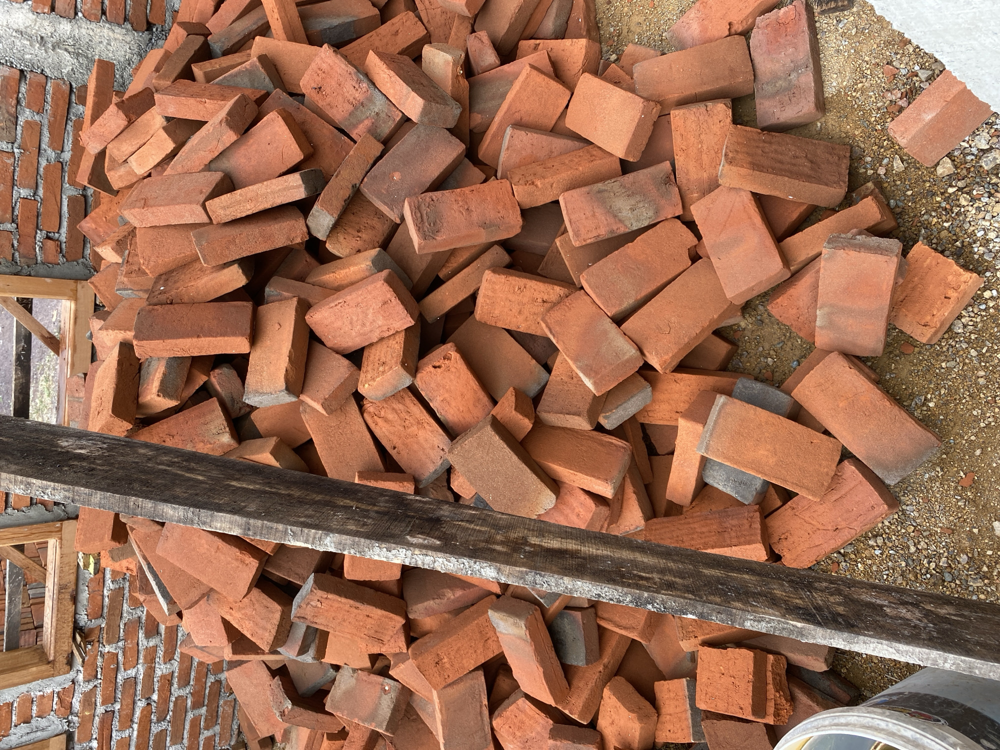 An outdoor, eye-level medium shot captures a pile of red bricks on a construction site. A weathered wooden plank runs vertically down the left side of the frame, partially obscuring the bricks. The bricks are of varying shades of red and orange, with some showing signs of wear and tear. They are piled haphazardly, creating a textured and uneven surface. In the background, a partially constructed brick wall is visible, along with wooden framing for a window or door opening. The ground beneath the bricks appears to be dirt and small stones. A white bucket with some dark liquid is partially visible in the bottom left corner. The overall scene suggests an active construction or renovation project.