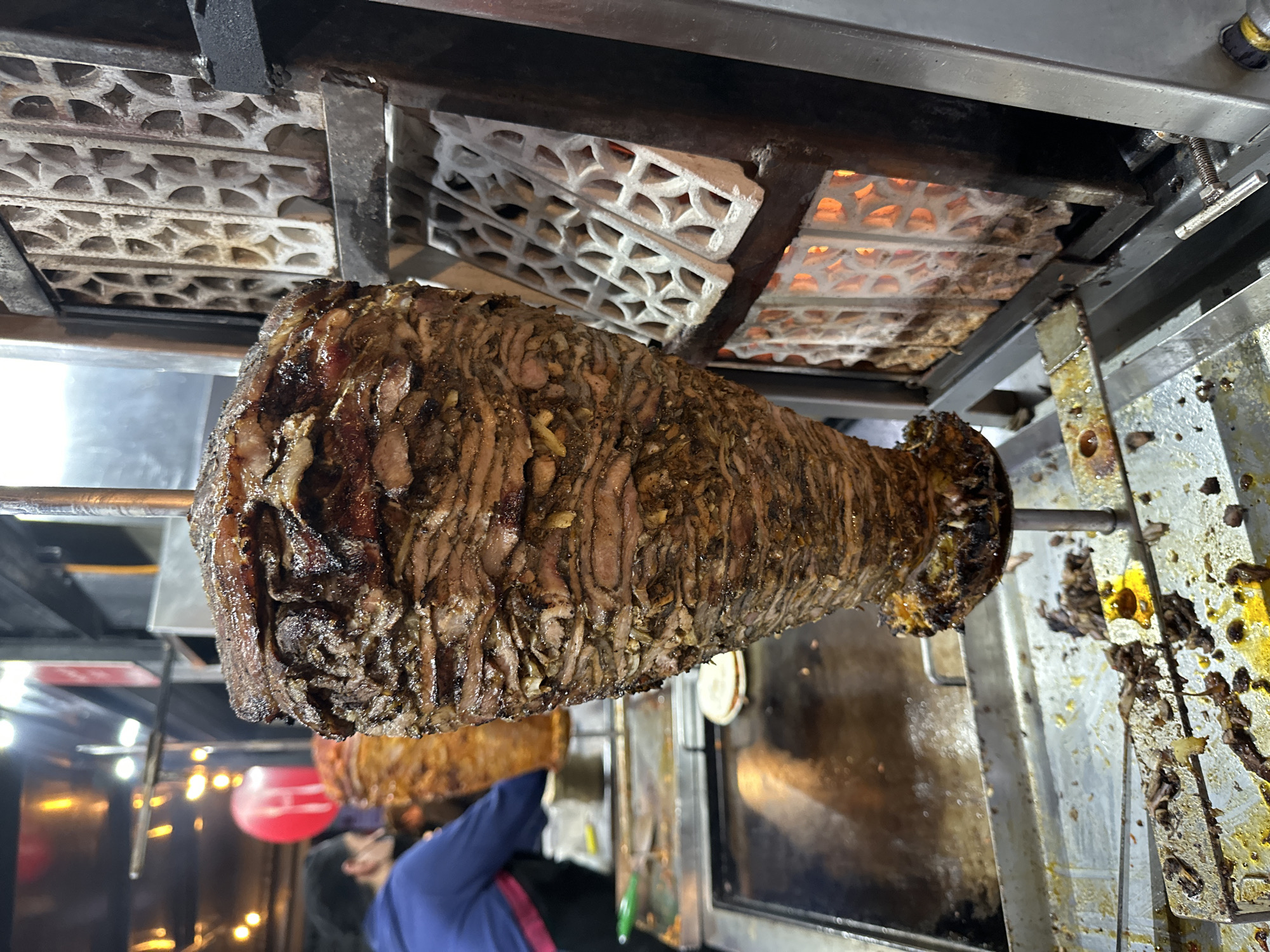A large, vertically rotating trompo of dark brown, seasoned meat, consistent with al pastor, is centrally positioned within a food preparation area in San Bernardino Tlaxcalancingo, Mexico. The meat, layered densely on a metal spit, exhibits visible cooked and charred surfaces. To the immediate right, a vertical heating element features glowing orange-red coils visible through white, geometric lattice grates, indicating active cooking. Beneath the trompo, a stainless steel drip tray collects rendered grease and meat particles. To the left, a dark griddle surface holds a single white tortilla. In the blurred background, another, smaller trompo of lighter-colored meat is present, along with a person wearing a blue hoodie and a dark face covering, standing behind a stainless steel counter. Overhead, multiple warm-toned lights illuminate the scene, suggesting a nighttime environment.