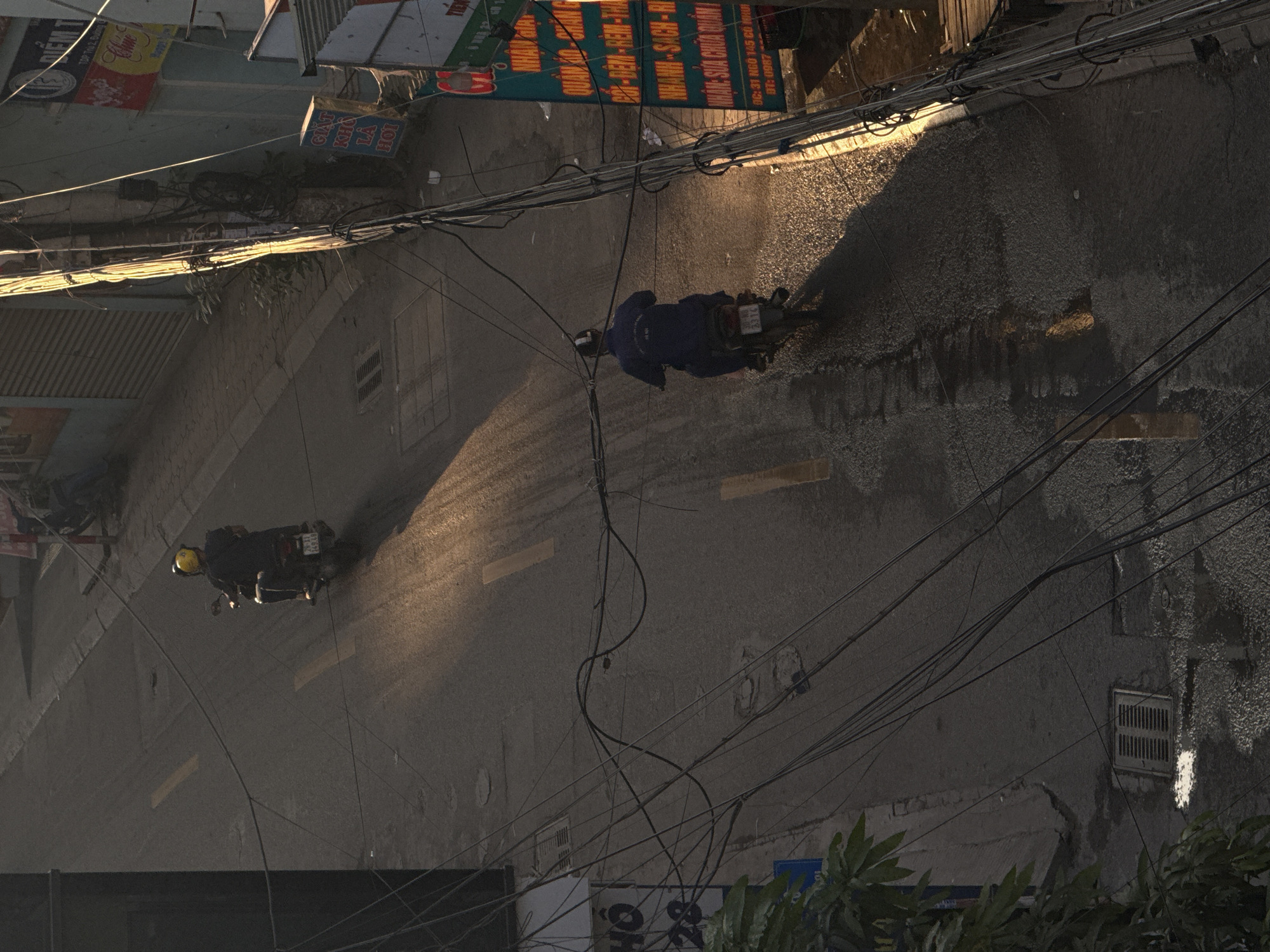 An eye-level, overhead shot captures a street in Hà Nội, Vietnam. Two people are riding mopeds on the street, with the person on the right wearing a dark blue outfit and the other wearing a yellow helmet. Power lines crisscross over the scene. Several buildings with advertisements and signs are on the left and top of the image.