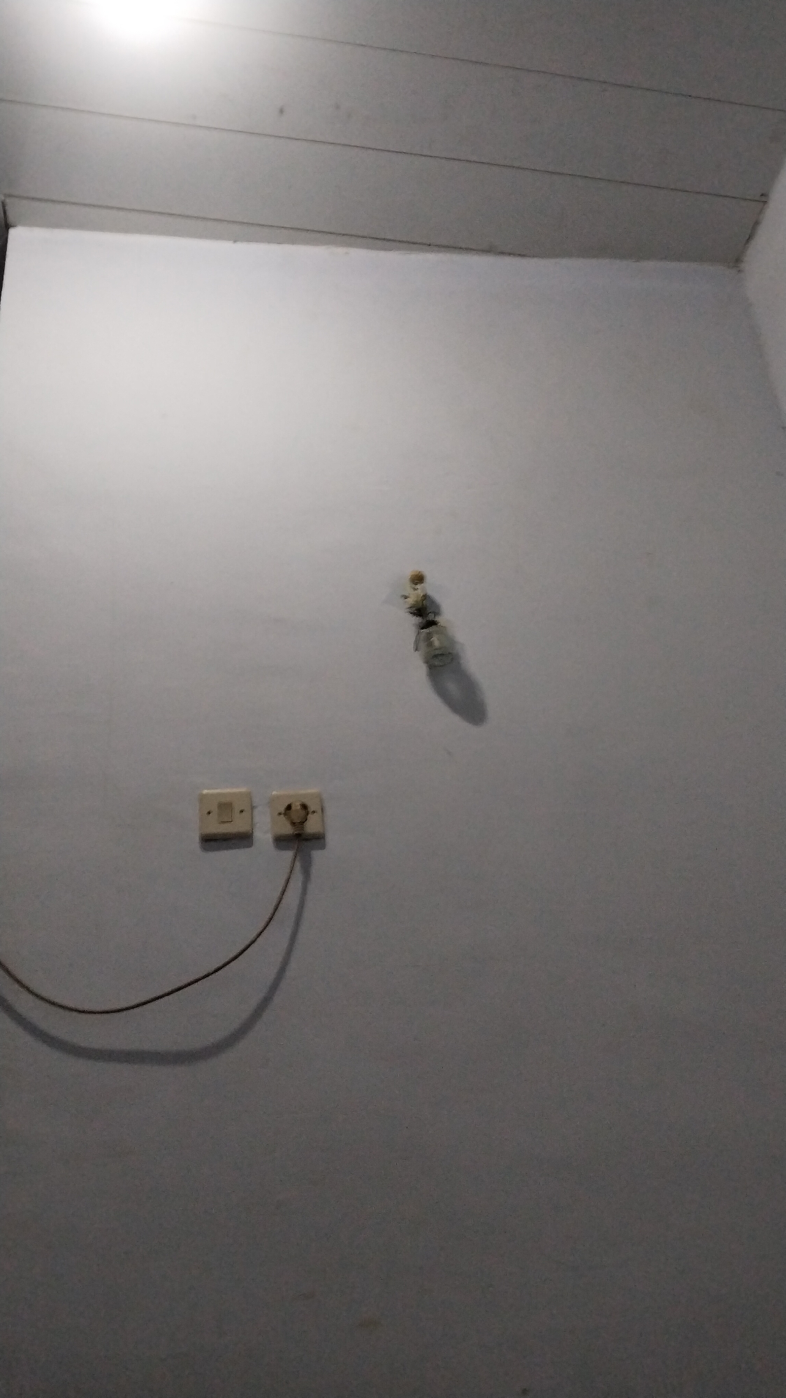 The image shows a white wall with a light fixture and two electrical outlets. One outlet is a standard two-prong outlet, while the other is a light switch. A wire is plugged into the outlet and hangs down the wall. The ceiling is visible at the top of the image, and it appears to be made of wood panels. The scene is dimly lit, and there is a shadow cast by the light fixture on the wall. The image appears to be taken indoors, in what could be a residential or commercial building. Based on the provided context, the location is the city of Banjarsari, Indonesia.