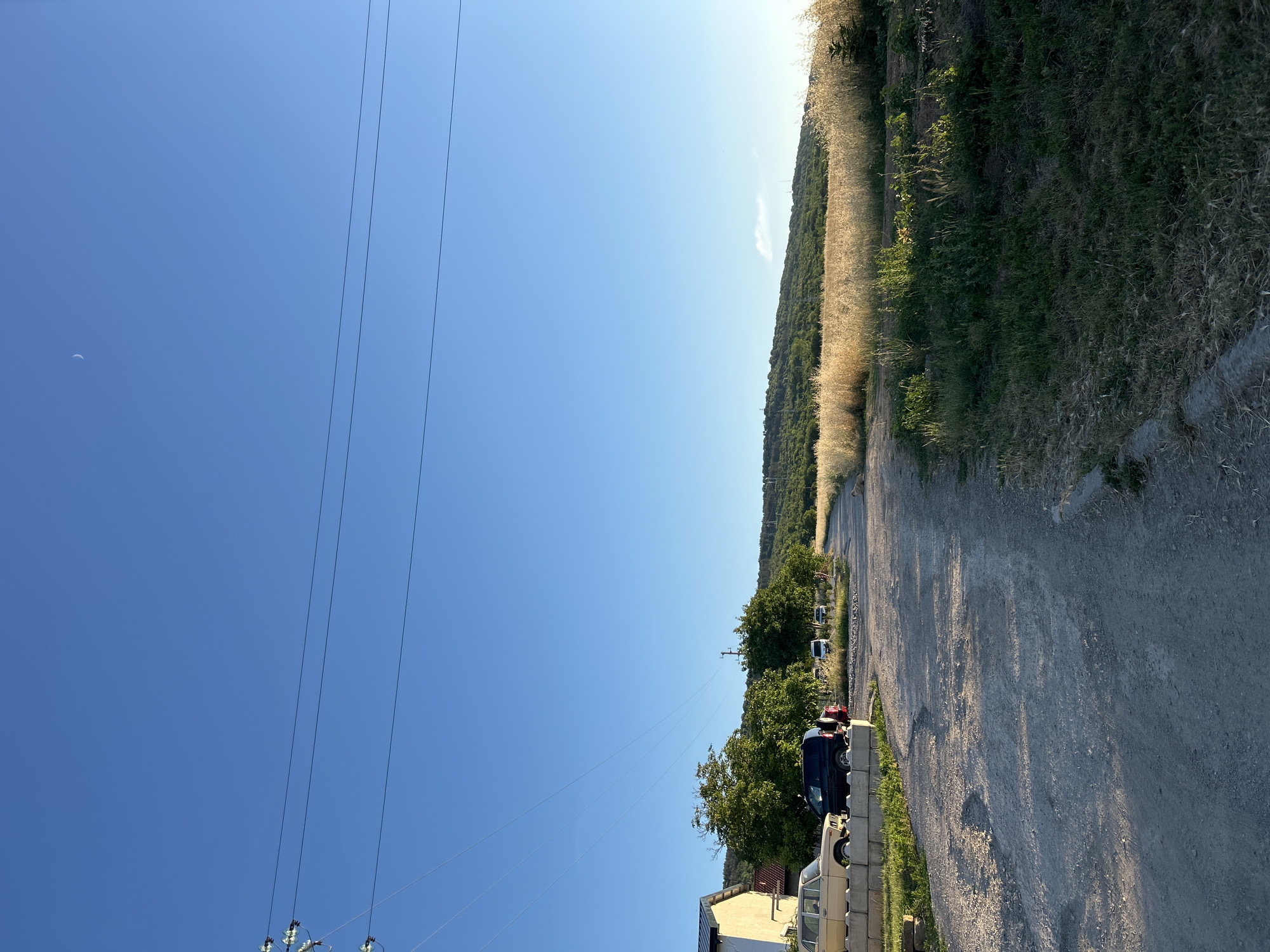 A dirt road winds through a rural landscape under a clear blue sky. On the left, a light-colored building with a white facade and a darker roof is visible, with a light-colored car parked in front of it. Next to the building, a dark-colored SUV is parked, and behind it, a few other vehicles are scattered along the road. Trees with lush green foliage dot the scene, providing shade. To the right of the road, a field of dry, golden grass stretches towards a gently sloping hill covered in green vegetation. The sun is casting long shadows, indicating it is either early morning or late afternoon. Power lines are strung across the sky, supported by utility poles. A faint crescent moon is visible in the upper right corner of the sky.