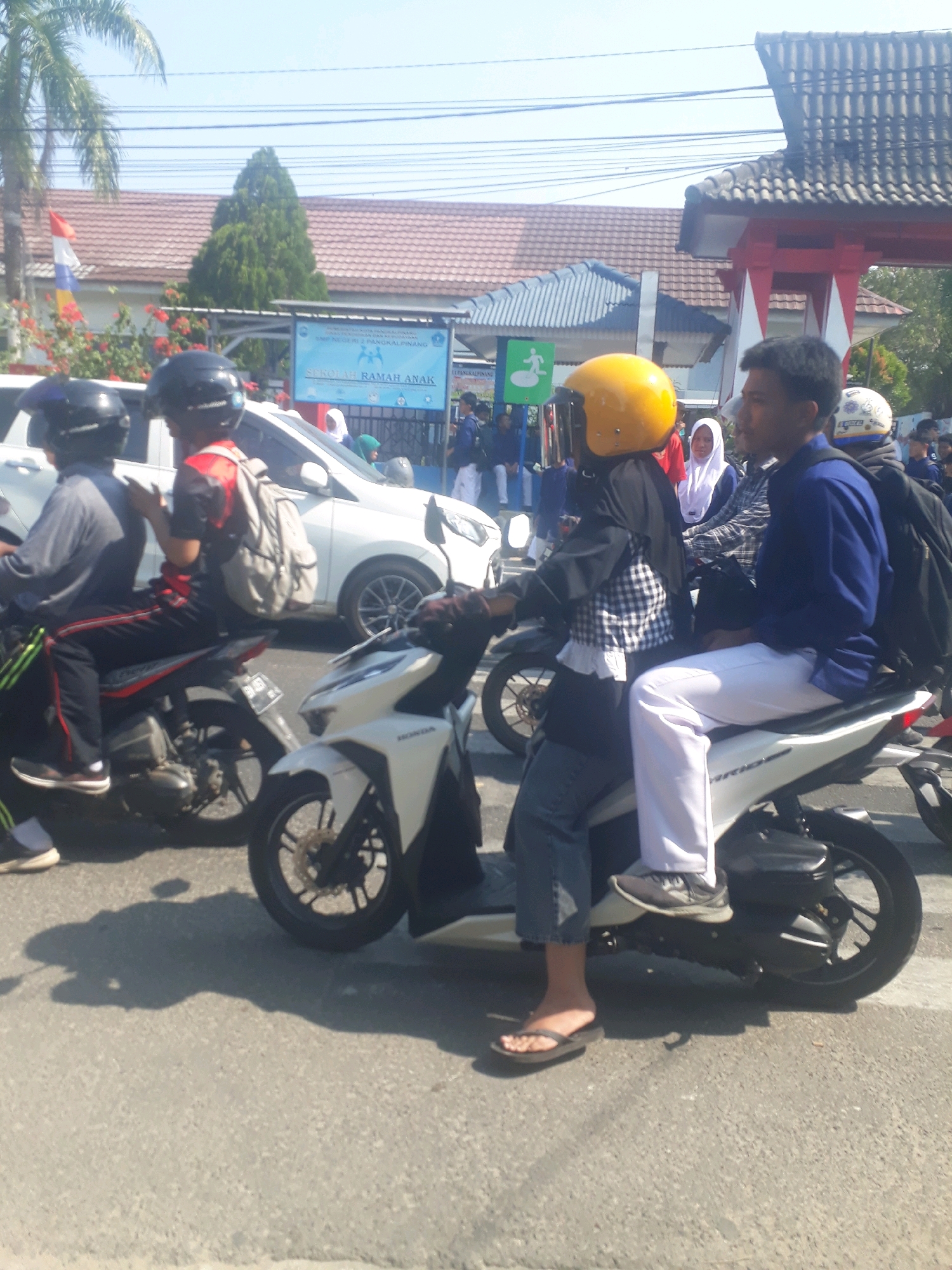 The image shows a sunny street scene in Pangkalpinang, Indonesia, with several people on motorcycles and a white car. In the foreground, two individuals are on a white motorcycle. The person in the back, wearing a yellow helmet and a black and white checkered shirt, is seated, while the person in the front, wearing a blue shirt and white pants, is also seated and looking to the right. Behind them, another person on a motorcycle wears a black helmet and a gray shirt. A white car is visible to the left. In the background, a building with a red and white facade and a tiled roof is present, along with trees and power lines. A blue sign with the text "SEKOLAH RAMAH ANAK" (Child-Friendly School) is also visible. The ground is paved with asphalt.