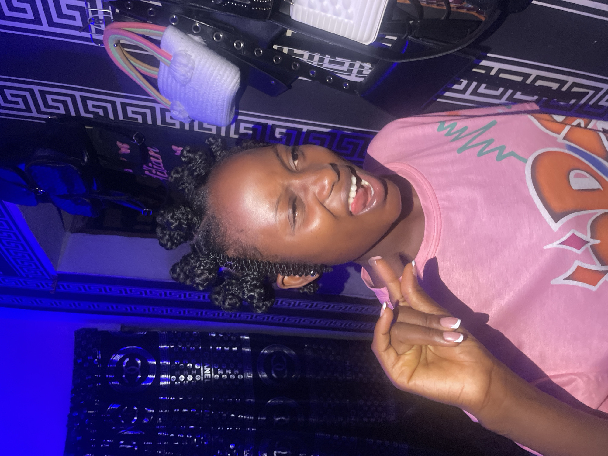 A woman with dark hair styled in twists is smiling with an open mouth, seemingly posing for a picture. She is wearing a pink t-shirt. Her left hand is visible, displaying a manicure with white tips. The background includes a black and white patterned surface with an object wrapped in a white cloth and secured with colorful cords on top of it. Below this area, there is a wall or a surface with a blue-toned ornate design. Part of the surface is adorned with a Chanel logo. The location appears to be indoors. There is not enough information to verify if the location is in the city of Lokoja, Nigeria.