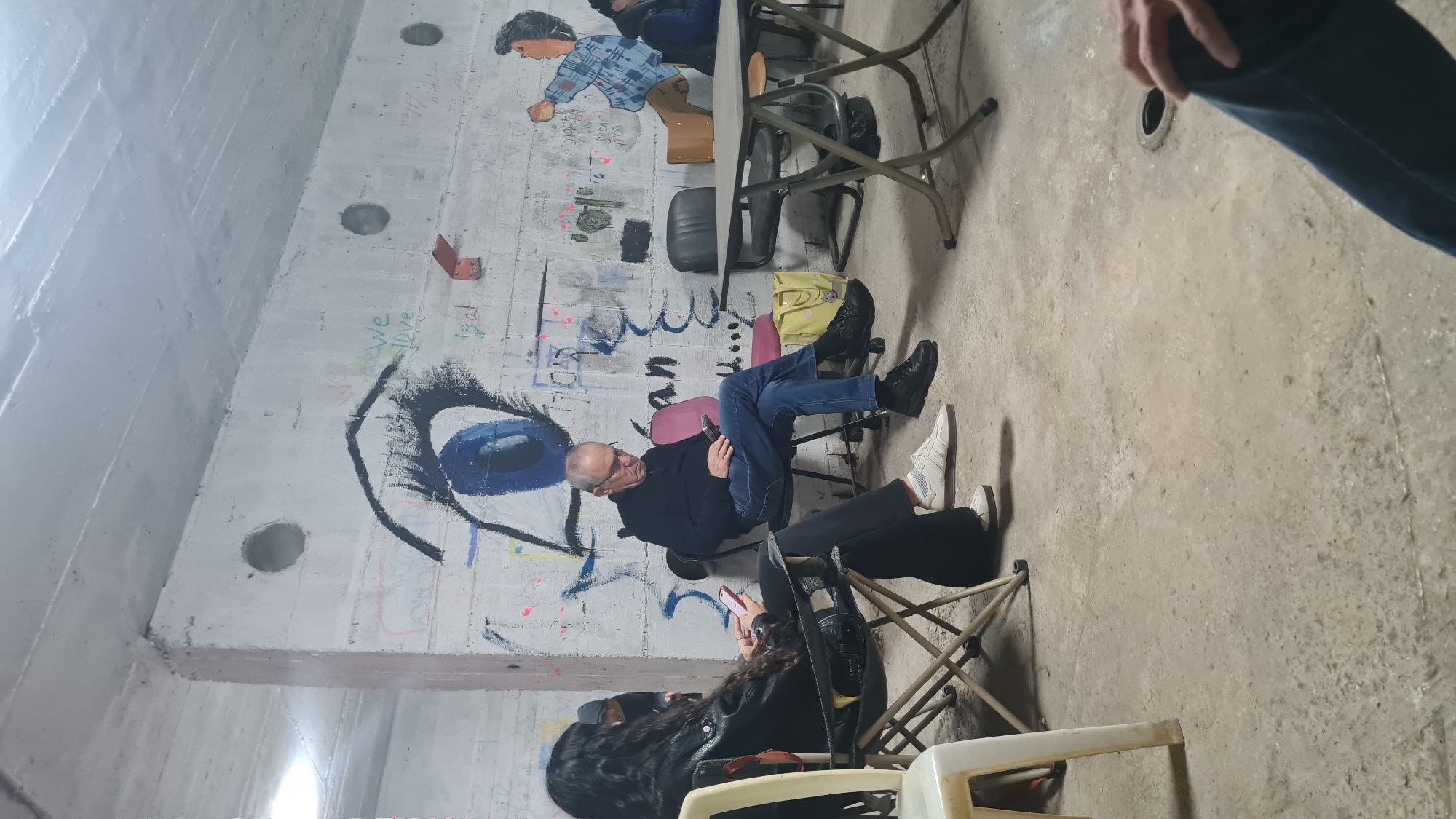 The photograph shows an interior space, likely a bunker or reinforced structure, with concrete walls covered in graffiti. The graffiti includes a large depiction of an eye and a stylized drawing of a person. Several people are seated on folding chairs, some looking at their phones. There are tables present. The location is potentially Rishon LeZion, Israel, but cannot be definitively determined solely from the image.