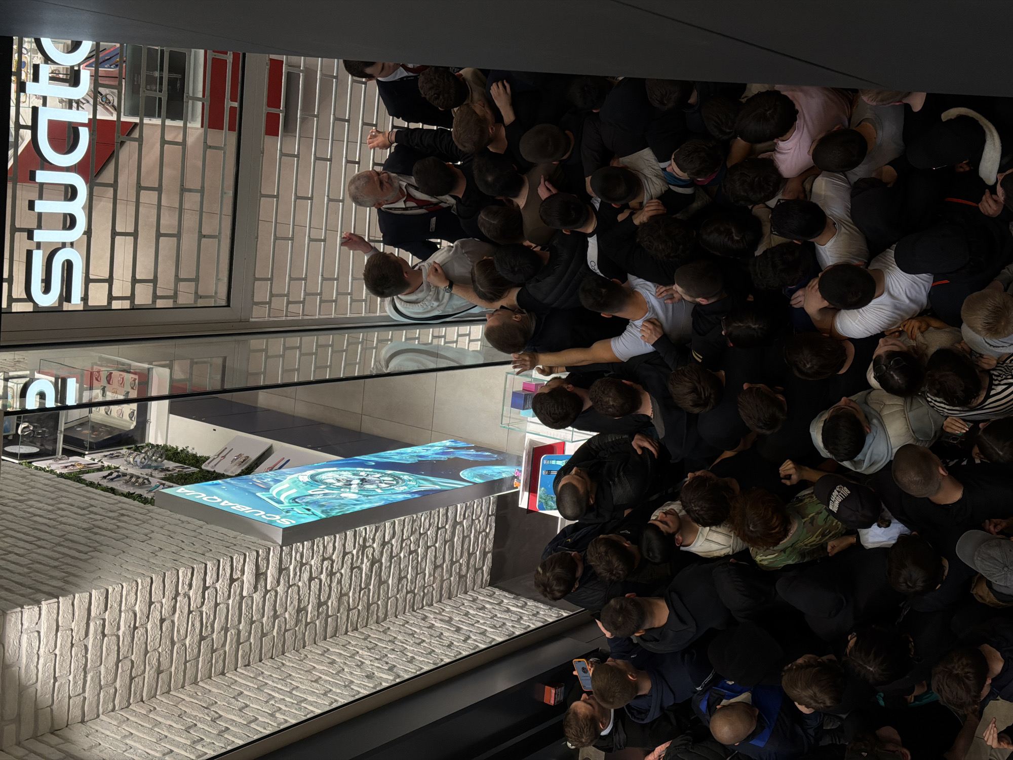 A high-angle shot shows a large crowd of people congregated outside a Swatch store. The store is identified by the prominent "Swatch" sign in block letters. The store is closed, as indicated by the metal security grille pulled down over the entrance. The crowd is densely packed and appears to be eagerly awaiting something, possibly the store opening. The crowd is predominantly composed of young adults and adults. The Swatch store is located in Belgrade, Serbia.
