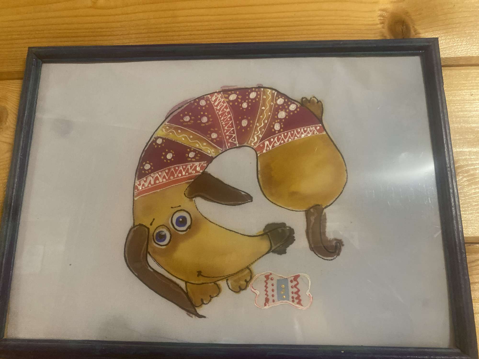 A framed painting of a cartoon dachshund dog is displayed against a light blue background. The dog is yellow with brown floppy ears and a curled brown tail. It is wearing a red and yellow patterned sweater that wraps around its body. The dog has large blue eyes with black pupils and a small smile. To the lower right of the dog, there is a small bone-shaped decoration with red and blue stitching. The painting is framed in a dark blue frame. The framed artwork is positioned on a wooden surface, possibly a wall or table, with visible wood grain. The background of the image is a wooden surface, likely a wall with horizontal planks of wood.