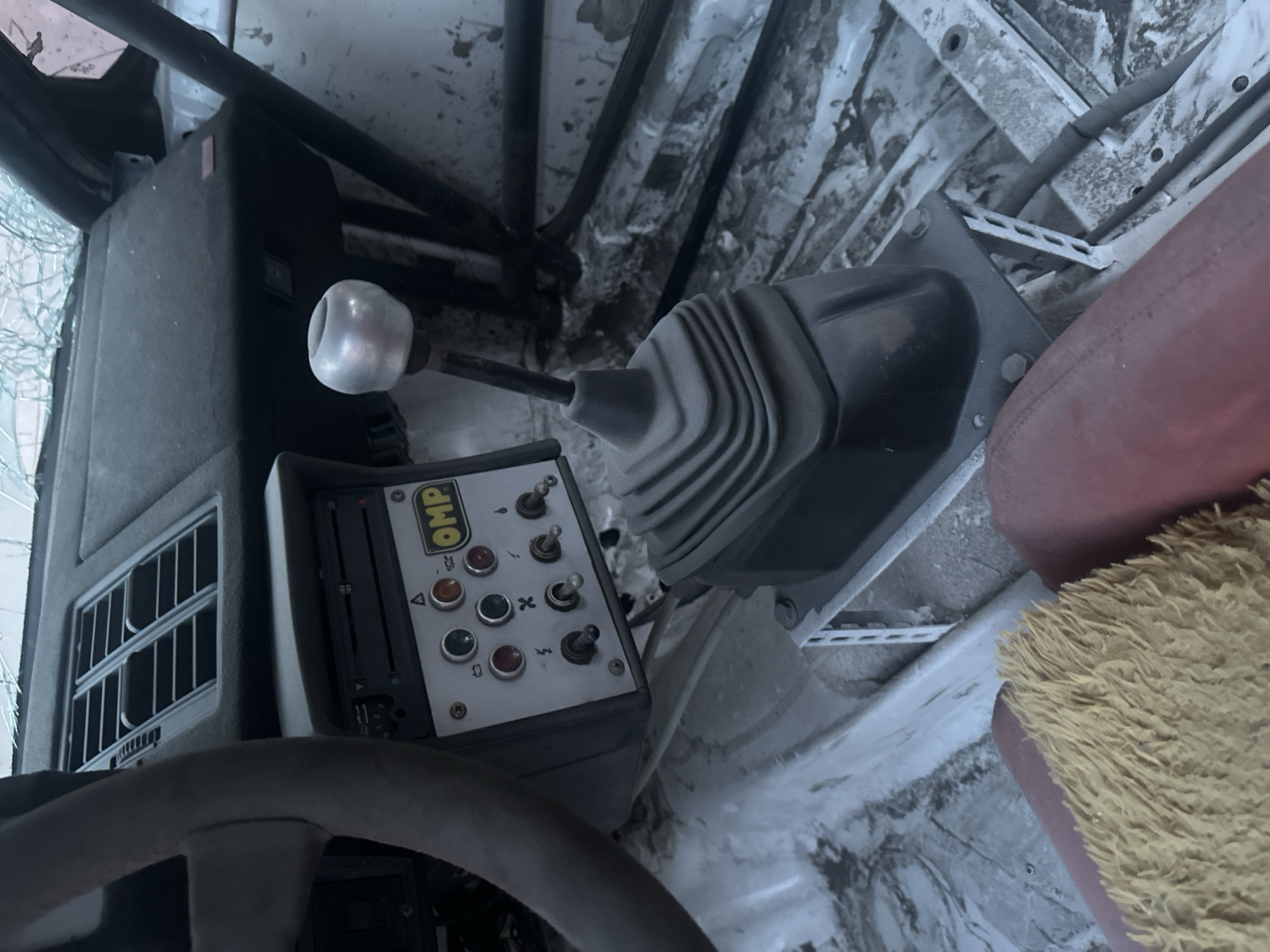 This image shows the interior of what appears to be a vehicle, possibly a race car or utility vehicle, given the presence of a manual gear shifter, a control panel with multiple switches and buttons, and a steering wheel. The overall setting is somewhat utilitarian and appears to be worn or used.
The central focus is on the gear shifter, which has a metallic, spherical knob. To the left of the shifter is a control panel with the "OMP" logo prominently displayed. This panel features several toggle switches and push buttons, some with indicator lights, suggesting control over various functions. A steering wheel is visible in the foreground, partially obscuring the view.
The interior surfaces appear to be made of textured plastic or similar materials, with some signs of wear and tear. The background shows structural elements of the vehicle, including metal framing and what looks like the vehicle's interior wall, which has a distressed, almost grimy texture. A red seat is visible on the right side of the frame, with a fuzzy yellow material in the bottom right corner, possibly a floor mat or seat cover.
There is no clear indication of people or a specific activity taking place, other than what can be inferred from the vehicle's controls. The lighting suggests it might be daytime, but there are no strong environmental cues like windows showing the outside. The location is identified as Comrat, Moldova. The visible text includes the "OMP" logo on the control panel, along with some symbols and labels on the switches and buttons, though these are not clearly legible.