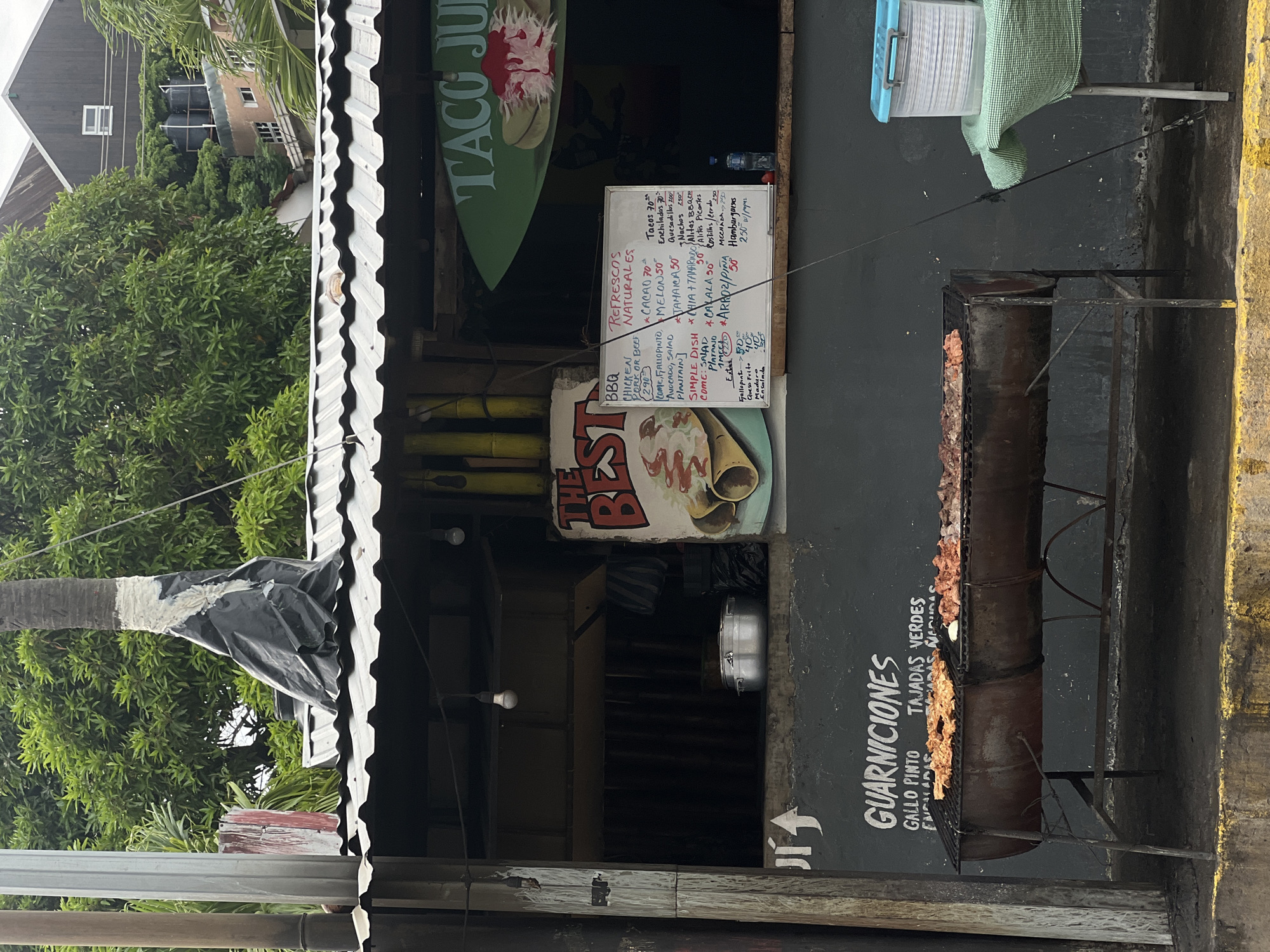 The image depicts a street-side food stand in San Juan del Sur, Nicaragua. The food stand features a corrugated metal roof with visible lighting fixtures. In front of the stand, a large grill is set up, loaded with various meats, possibly pork and chicken. To the left of the grill, text on the wall reads "GUARNICIONES GALLO PINTO TAJADAS VERDES ENCILADAS NACHOS". A menu board is also visible, listing items like "BBQ CHICKEN PORK OR BEEF", "REFRESCOS NATURALES", and various types of tacos and burgers, with prices indicated. A cooler filled with what appear to be stacked plates or containers sits on a table to the right of the grill. The background shows lush green trees and buildings, characteristic of the tropical environment of San Juan del Sur. The overall scene suggests a vibrant, open-air eatery preparing to serve customers.